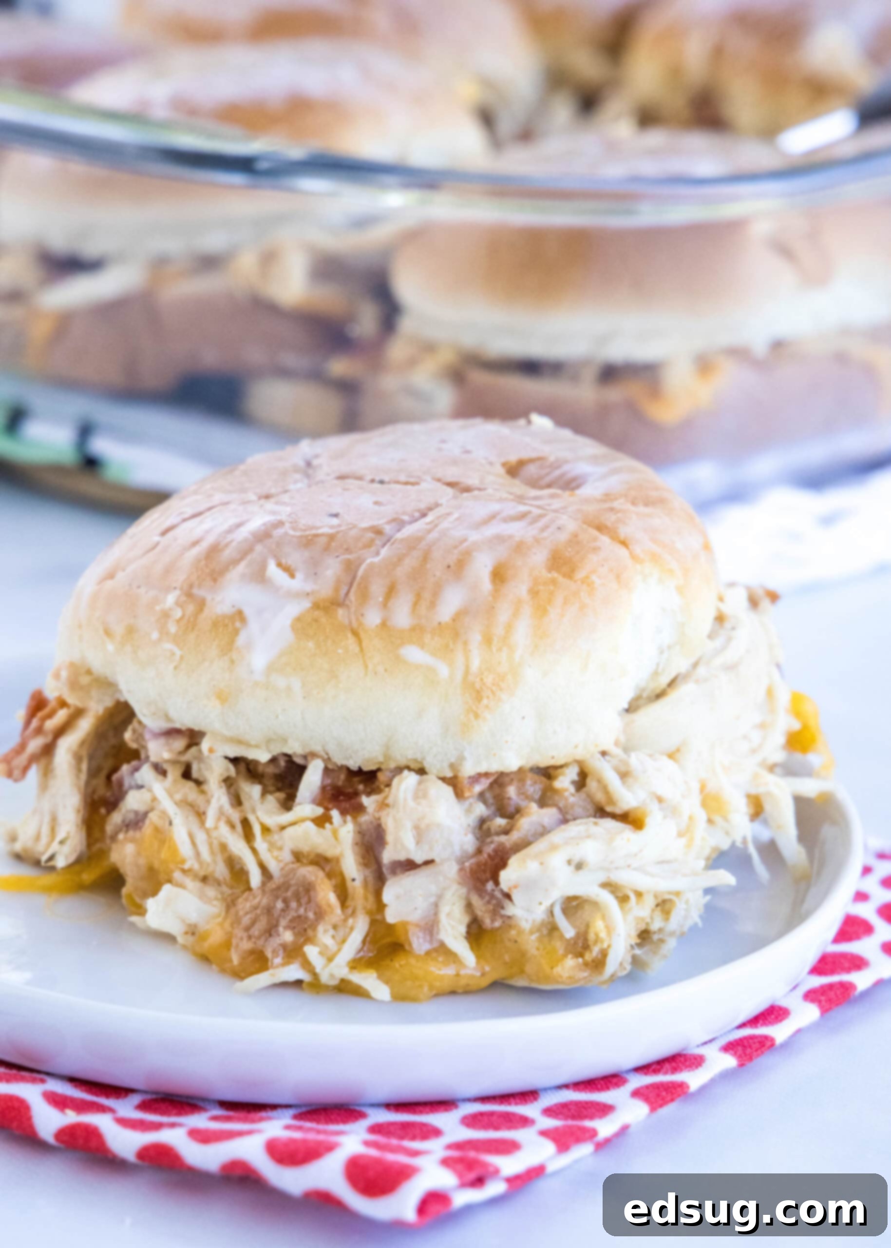 Make my easy chicken bacon ranch sliders with juicy shredded chicken, bacon, and creamy ranch dressing between sweet, fluffy Hawaiian rolls! A chicken bacon ranch slider on a plate with more sliders in a glass baking dish in the background.