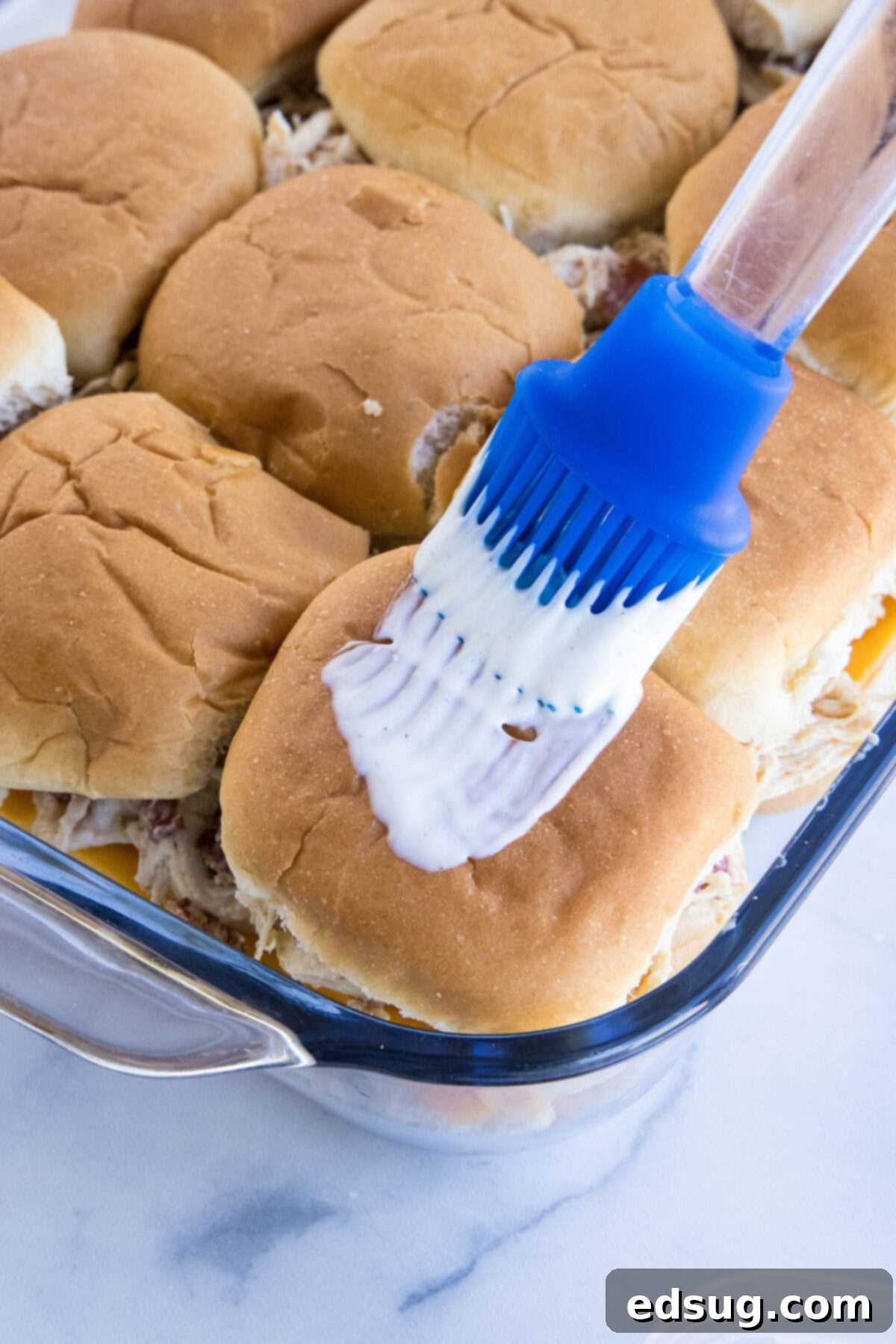Make my easy chicken bacon ranch sliders with juicy shredded chicken, bacon, and creamy ranch dressing between sweet, fluffy Hawaiian rolls! A rubber basting brush brushing ranch dressing over the tops of the slider rolls in a baking dish.