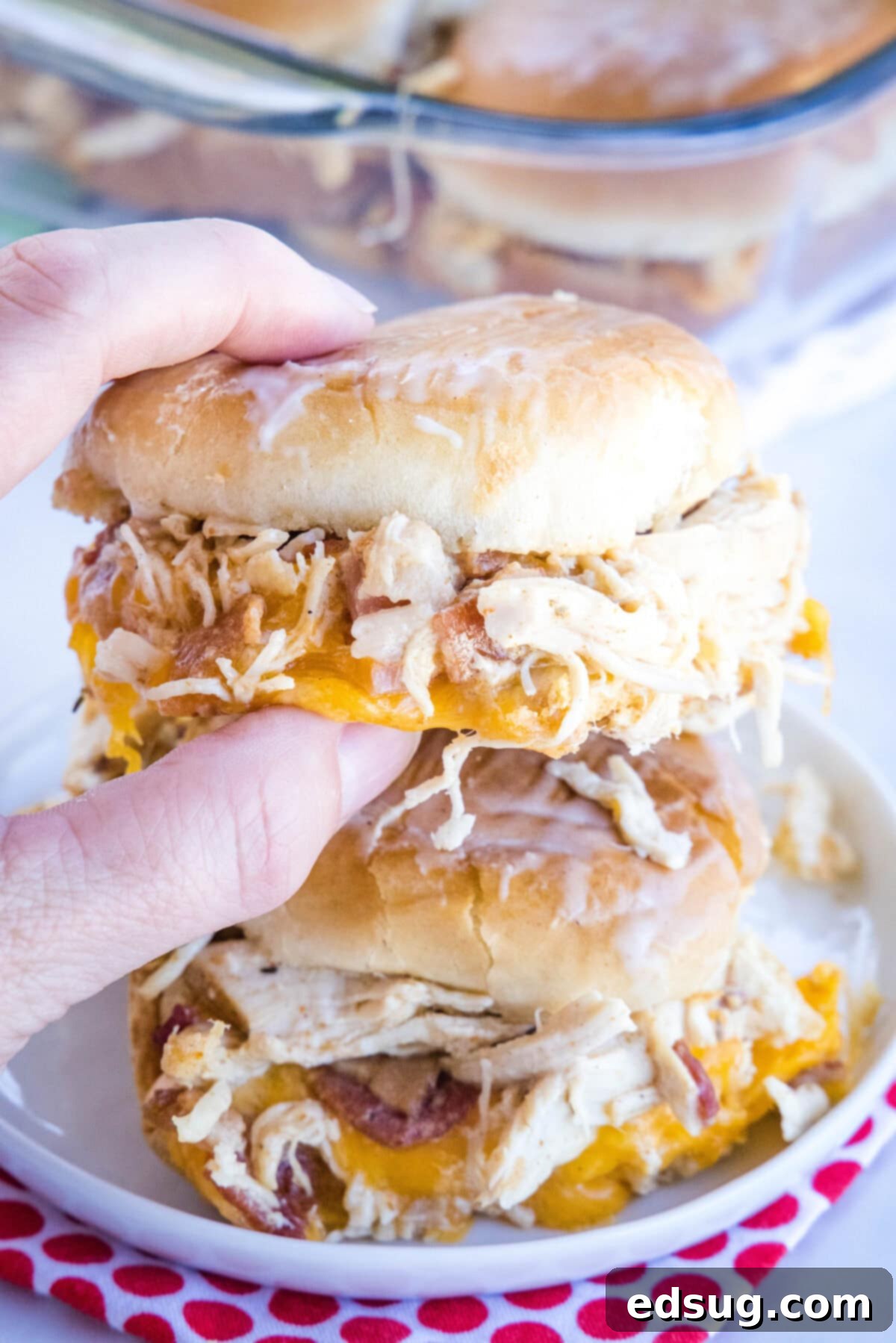Make my easy chicken bacon ranch sliders with juicy shredded chicken, bacon, and creamy ranch dressing between sweet, fluffy Hawaiian rolls! Close up of a hand holding up a chicken bacon ranch slider with a second slider on a plate in the background.