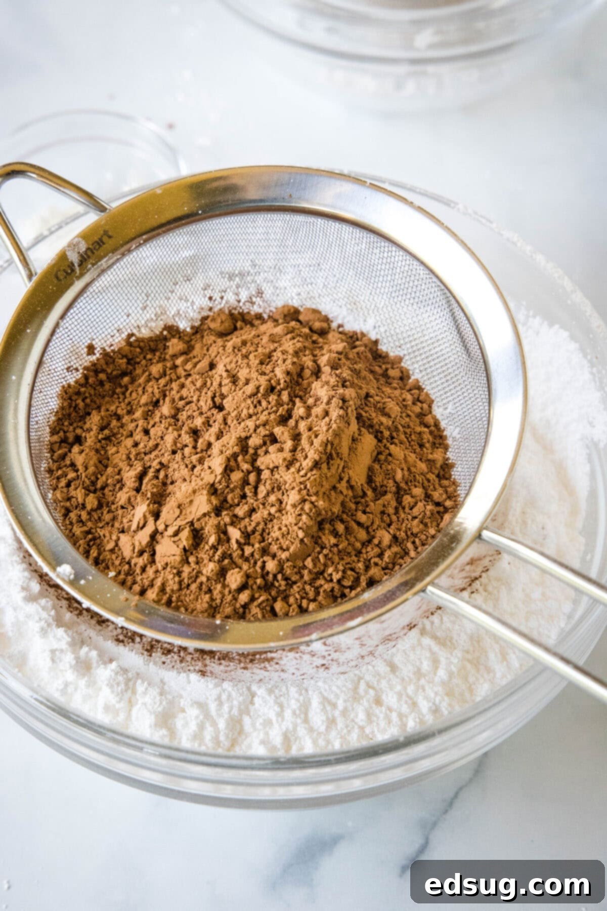 Cocoa powder in a sifter held over sugar in a glass bowl.