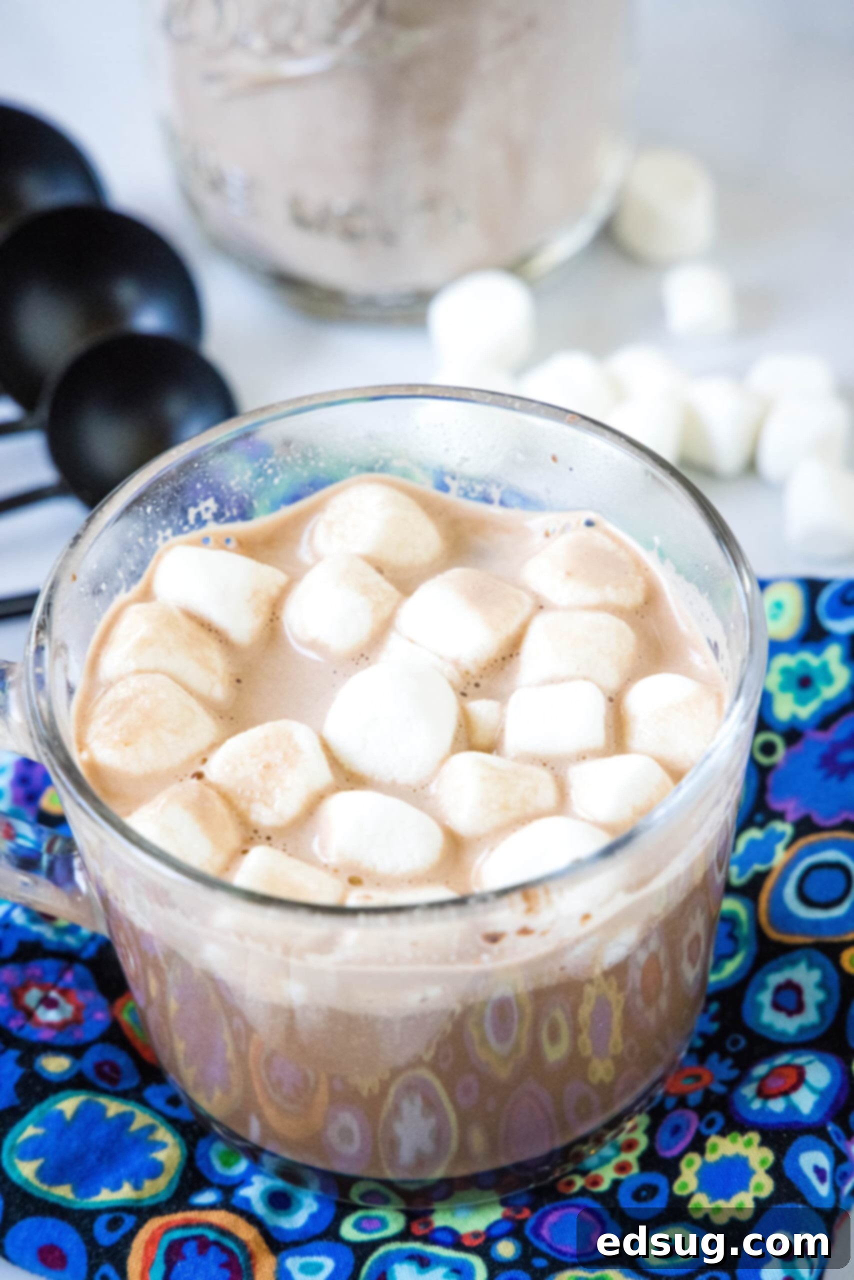 Hot chocolate in a clear mug topped with mini marshmallows, with more mini marshmallows and a jar of hot chocolate mix in the background.