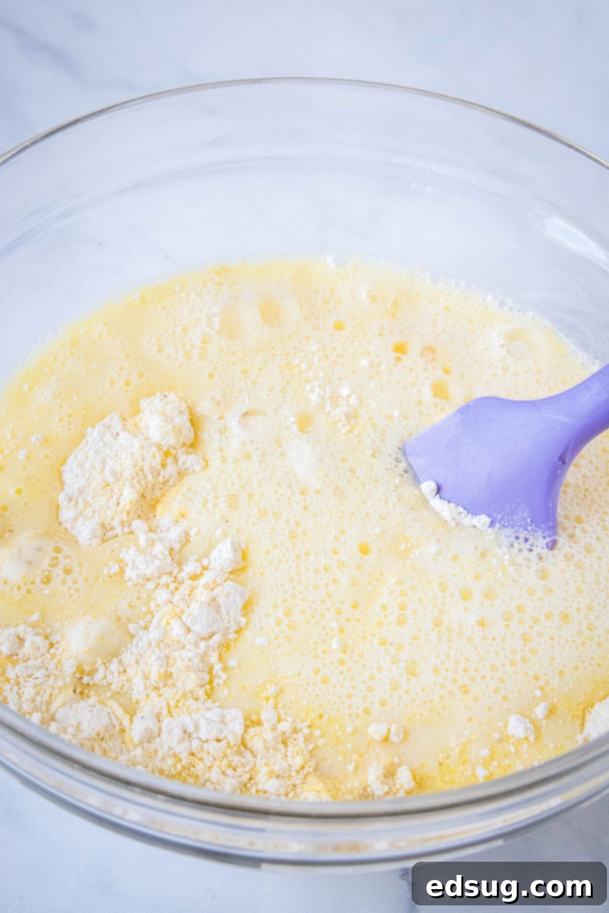 Combine your dry and wet ingredients carefully to form the perfect cornmeal pancake batter. The process of mixing cornmeal pancake batter in a clear glass bowl, using a vibrant purple spatula, showcasing the gentle combining of ingredients.