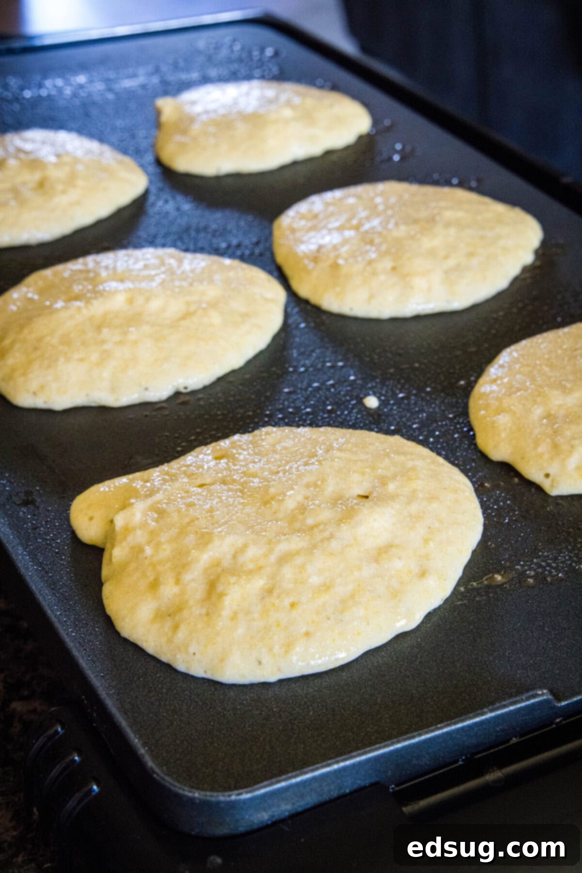 Watch for bubbles on the surface as your cornmeal pancakes cook to golden perfection on the griddle. Golden brown cornmeal pancake batter actively cooking and bubbling on a hot griddle, indicating readiness for flipping.