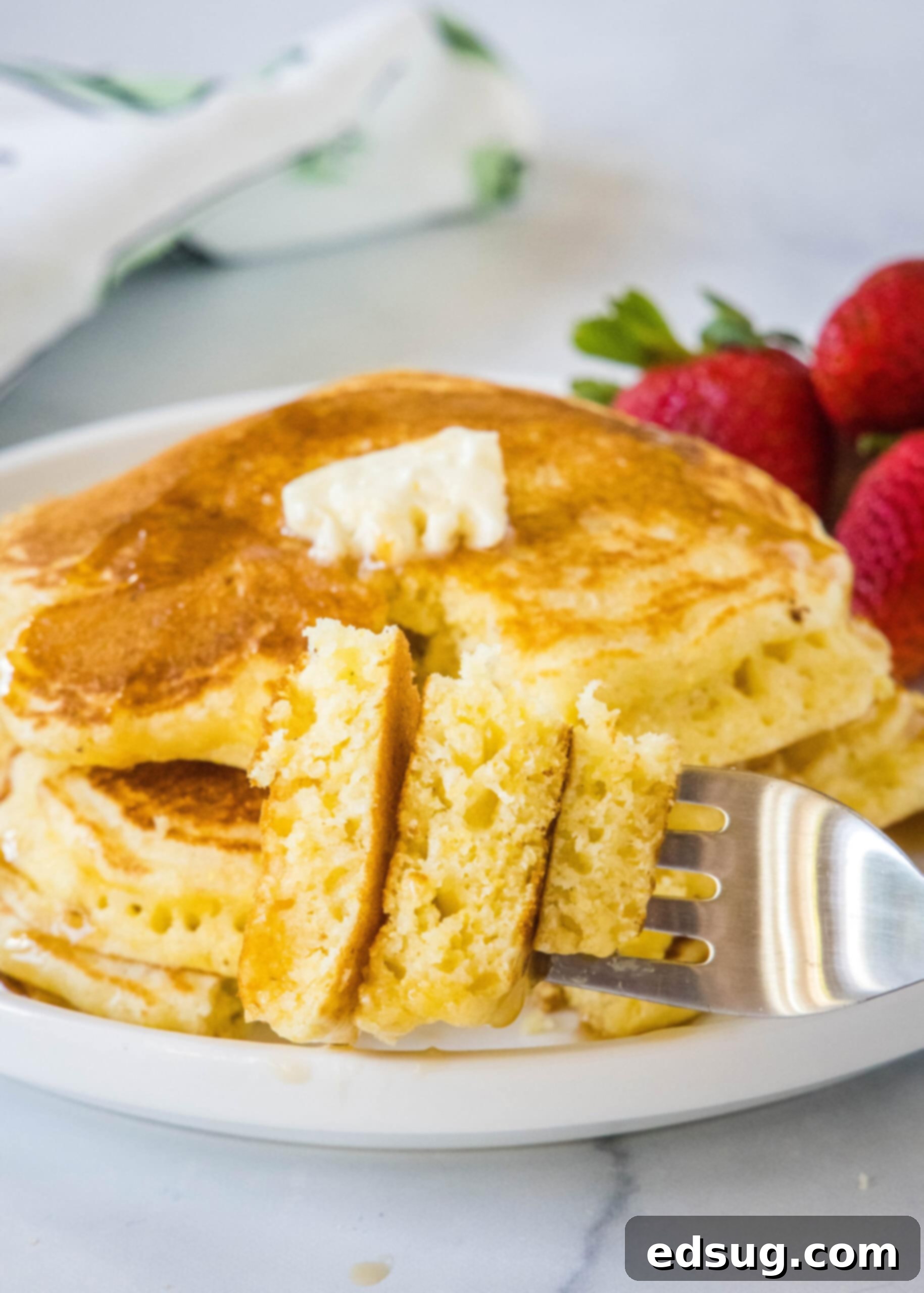 Savor a perfect bite of fluffy cornmeal pancake, showcasing its tender texture and golden hue. A fork piercing a fluffy cornmeal pancake, holding a perfect bite, with a stack of syrup-drizzled pancakes in the background on a white plate.