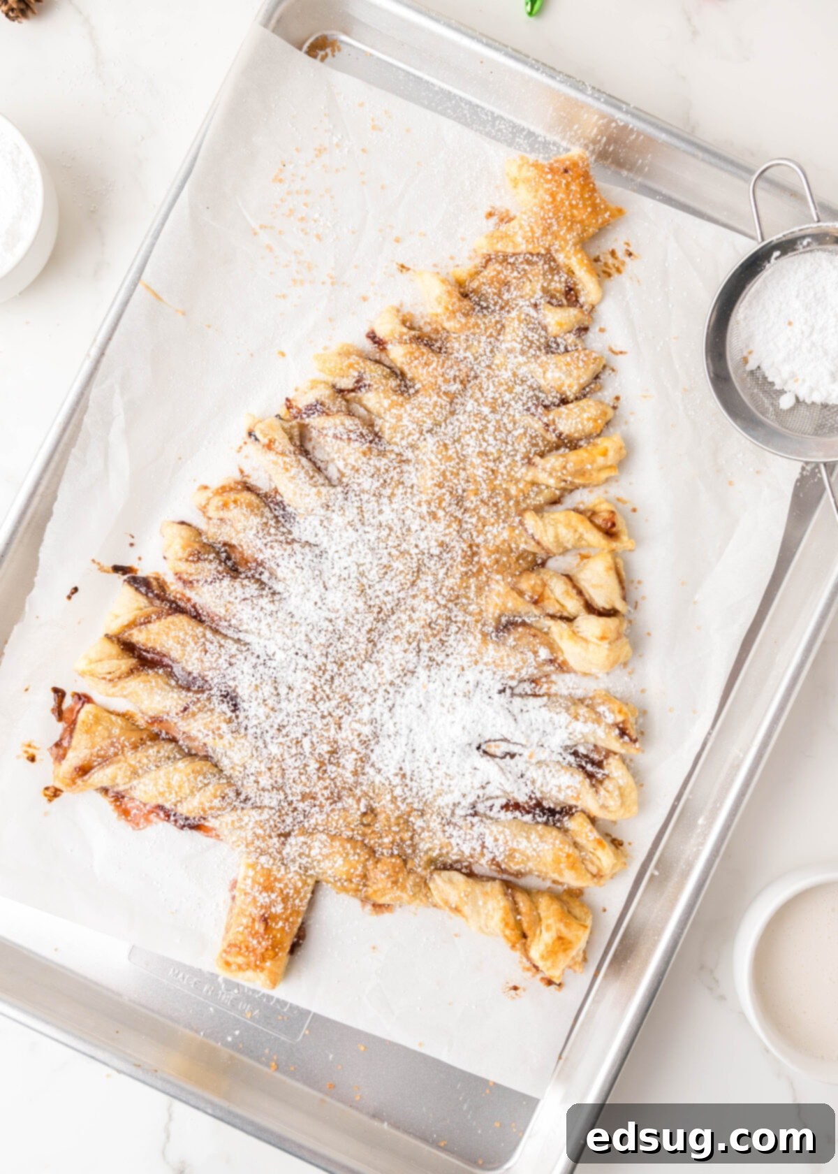 Holiday Swirl Pastry Tree 2 Powdered sugar delicately sprinkled over a golden-brown puff pastry Christmas tree, beautifully arranged on a baking sheet, ready to be served as a delightful holiday dessert.