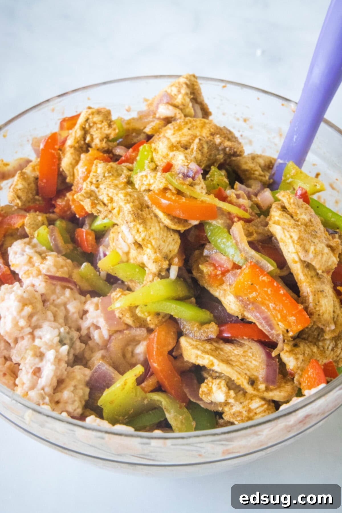 This chicken fajita casserole is an easy dinner packed with juicy chicken and rice, with sweet bell peppers, fajita seasoning, and cheese! Sautéed chicken, bell peppers, and onions are added to the rice, sour cream, and salsa mixture in a large glass bowl, ready to be stirred.