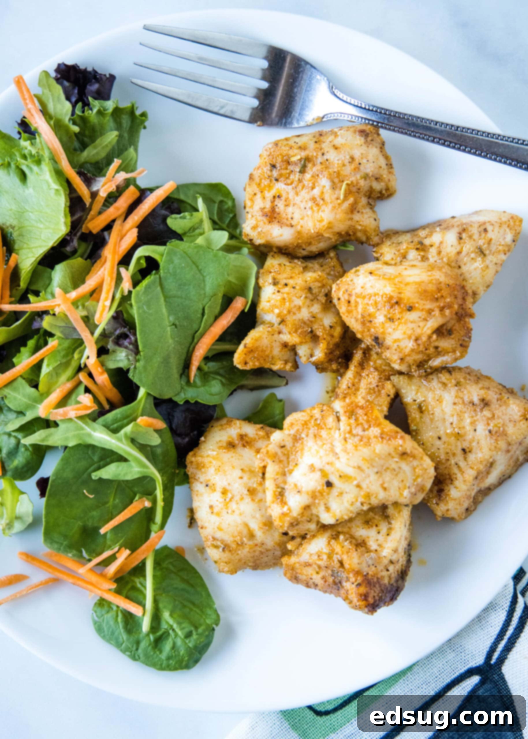 Baked chicken bites next to green salad and a fork on a plate. A plate featuring a generous serving of baked chicken bites accompanied by a fresh green salad and a fork, ready for a delicious meal.