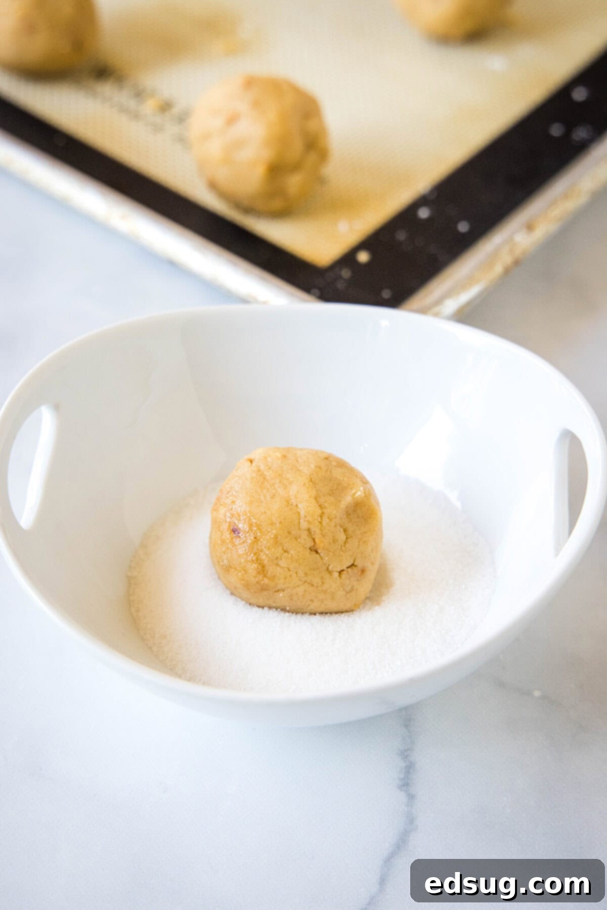 A ball of peanut butter cookie dough being rolled gently in a small bowl filled with shimmering granulated sugar.