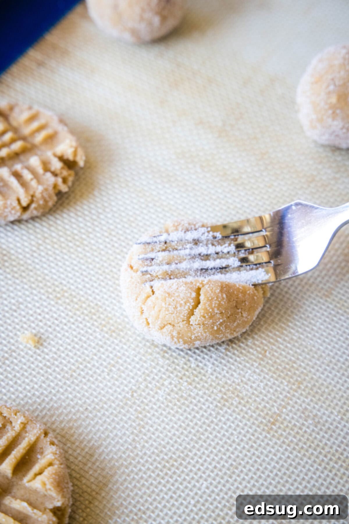 A pair of forks pressing down on soft peanut butter cookie dough balls, creating the iconic criss-cross pattern on a lined baking sheet.