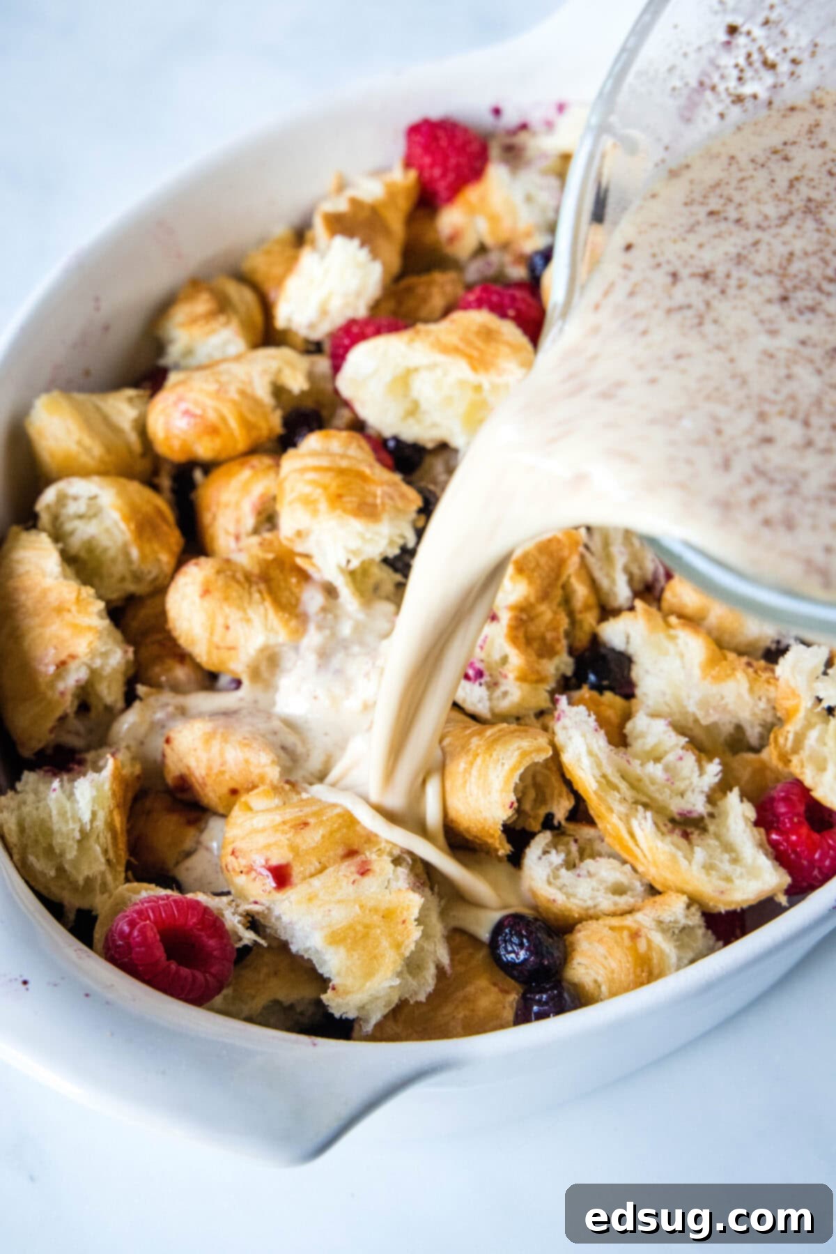 This overnight croissant baked French toast has buttery croissants layered in a rich, spiced custard with berries. It's prepped in minutes! Custard being poured over croissant pieces and fresh berries in an oval casserole dish, preparing for an overnight soak.