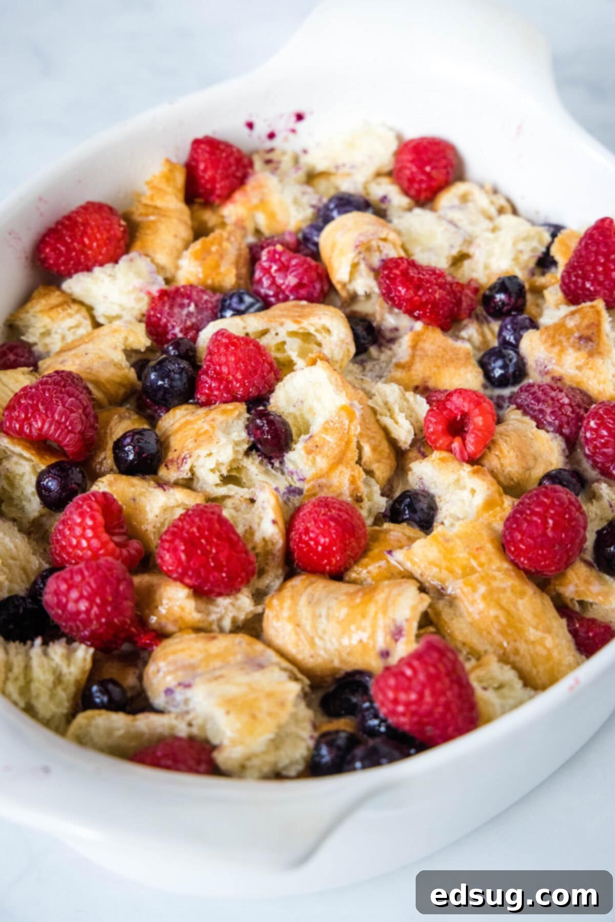 This overnight croissant baked French toast has buttery croissants layered in a rich, spiced custard with berries. It's prepped in minutes! Croissant pieces and fresh berries thoroughly soaked in egg custard inside an oval casserole dish, ready for refrigeration.