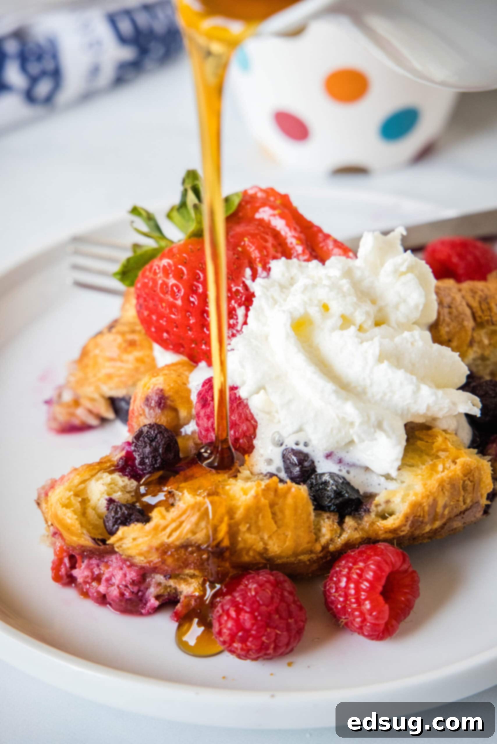 This overnight croissant baked French toast has buttery croissants layered in a rich, spiced custard with berries. It's prepped in minutes! Maple syrup being drizzled generously over a serving of croissant baked French toast, garnished with whipped cream and fresh berries.