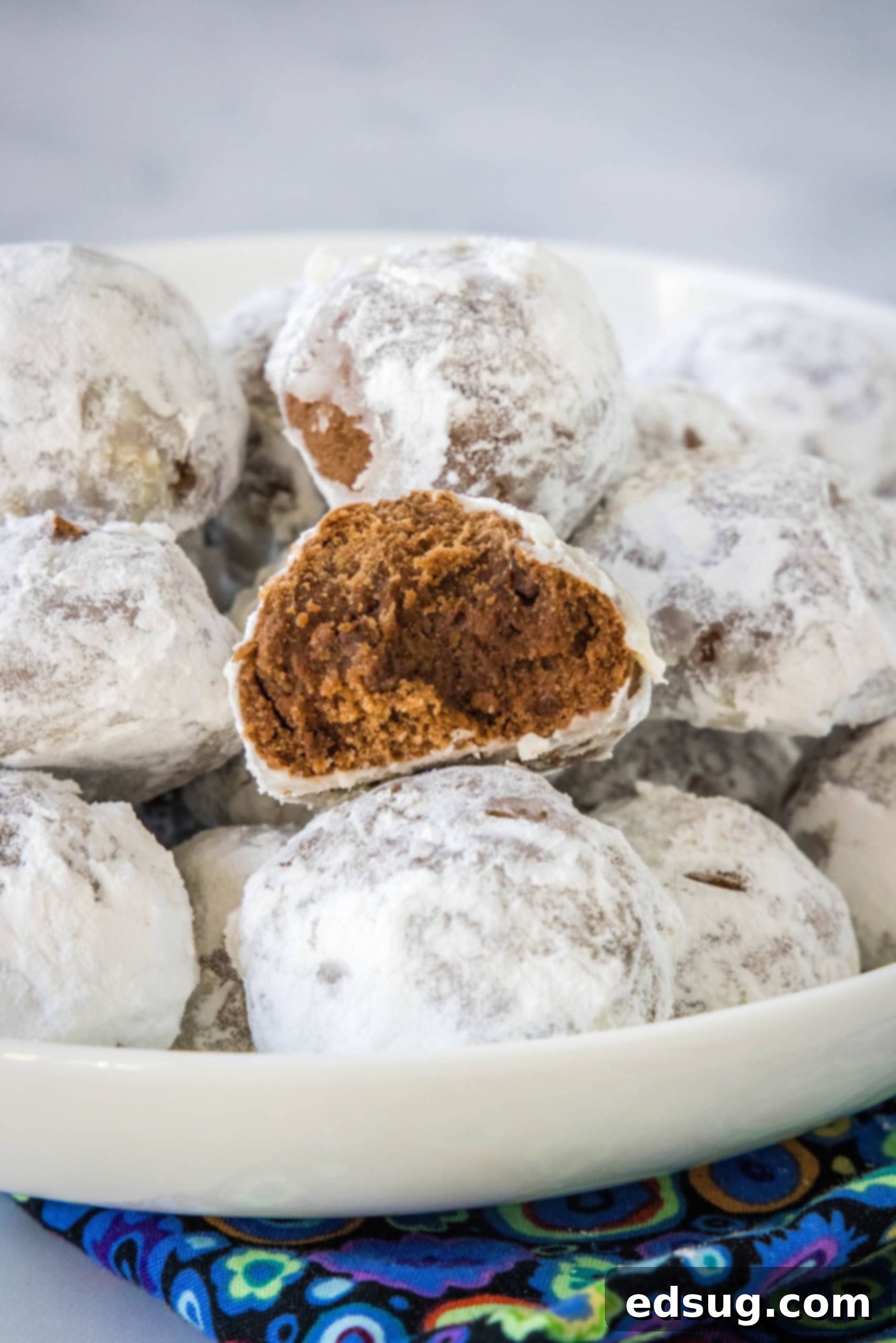 Chocolate snowball cookies are a fun twist on Mexican wedding cookies! These soft chocolate cookies are quick to make and irresistibly fudgy. Chocolate snowball cookies stacked on a white platter with a bite missing from one cookie, showcasing its fudgy interior.