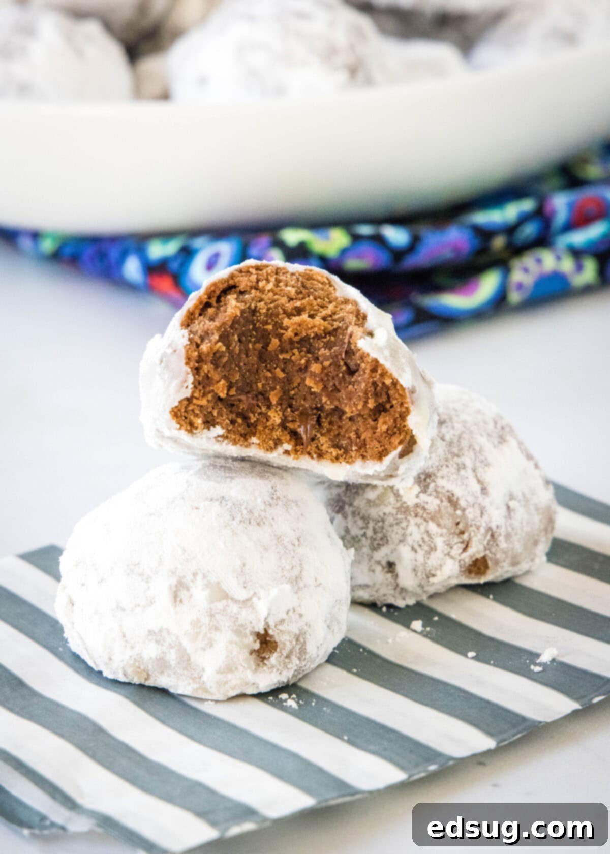 Chocolate snowball cookies are a fun twist on Mexican wedding cookies! These soft chocolate cookies are quick to make and irresistibly fudgy. Three chocolate snowball cookies stacked on a striped napkin, with a bite missing from the top cookie revealing its soft, fudgy interior.