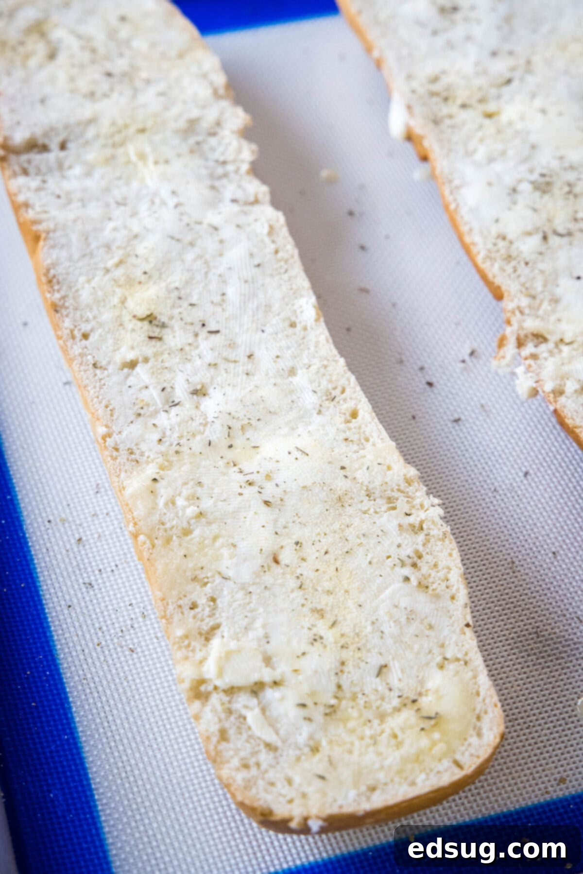 Prepare your French bread halves by spreading them generously with butter and sprinkling on fragrant seasonings. French bread halves spread with garlic butter on a baking sheet, ready for the oven.