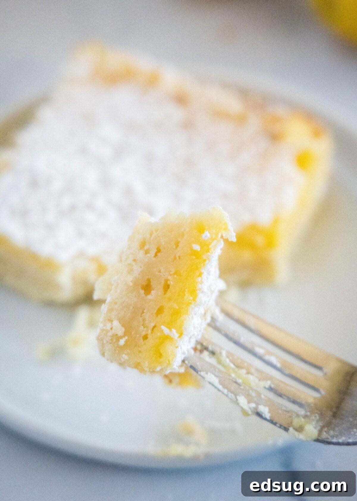 A bite of a lemon bar on a fork, with a full lemon bar on a plate in the background, showing the crisp crust and soft filling