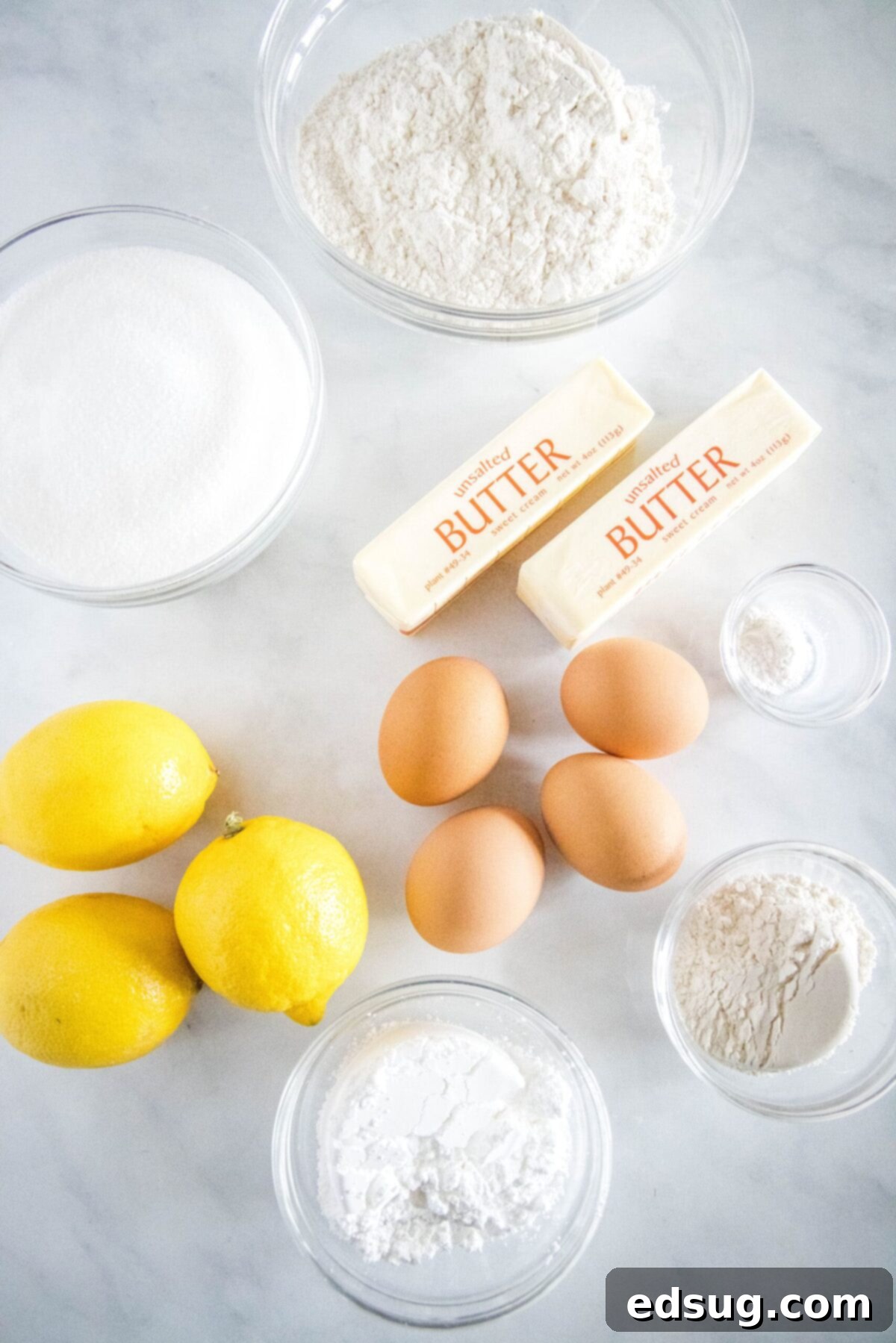 Overhead view of all the fresh ingredients needed for homemade lemon bars: a large bowl of all-purpose flour, a smaller bowl of sifted powdered sugar, a bowl of granulated sugar, a small bowl of baking powder, two sticks of unsalted butter, four fresh eggs, and three bright yellow lemons