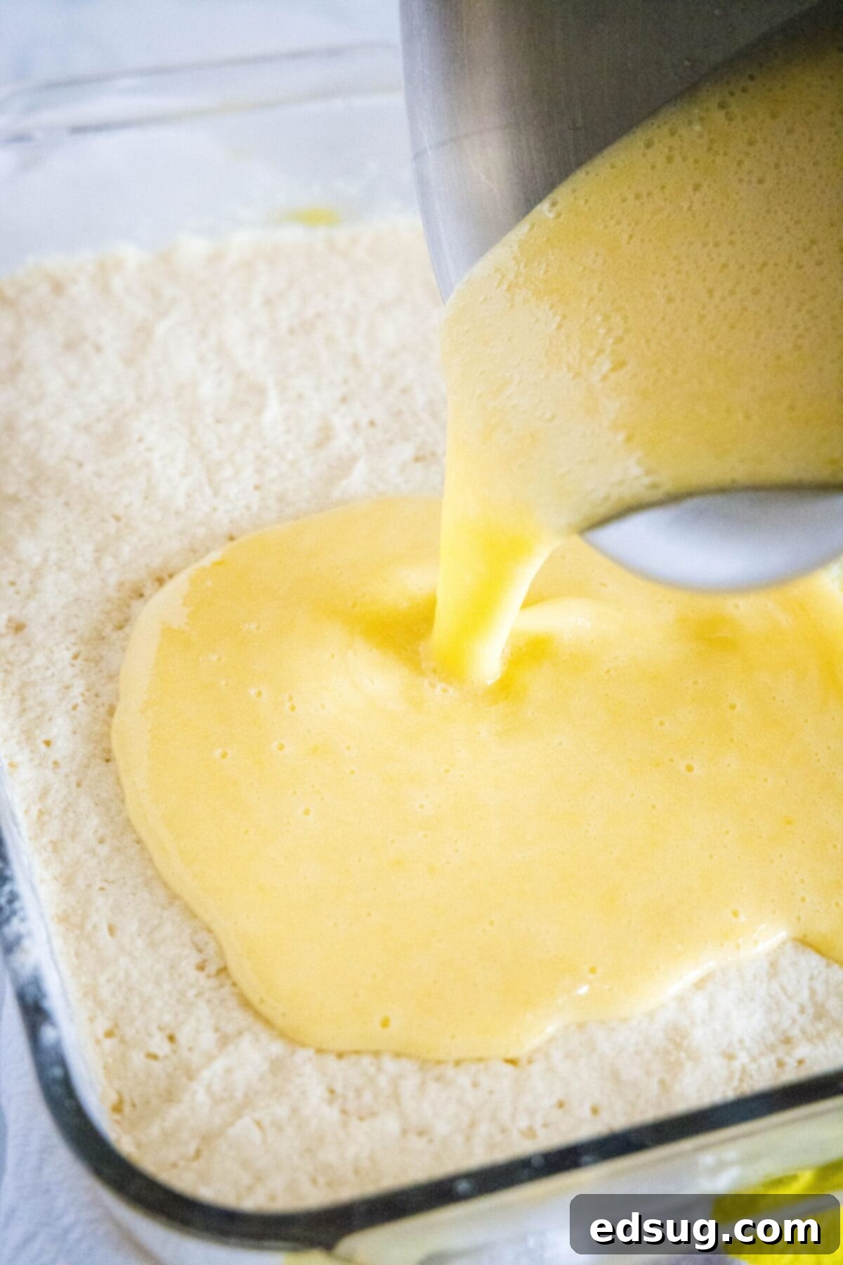 Vibrant yellow lemon filling being smoothly poured over the golden-brown shortbread crust in a baking pan