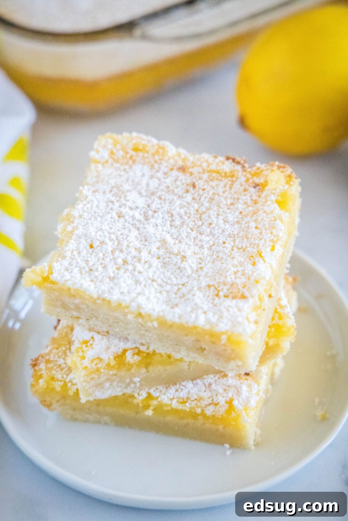 Three perfectly cut square lemon bars stacked neatly on a white plate, next to a whole fresh lemon