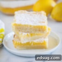 Three square lemon bars stacked on a plate with more bars and lemons behind them