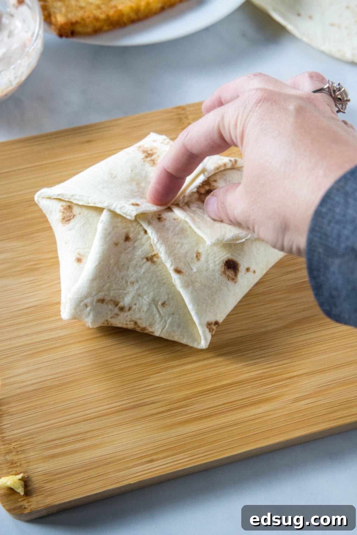 A hand expertly folding the edges of a large flour tortilla inward to form a breakfast crunchwrap on a wooden cutting board.