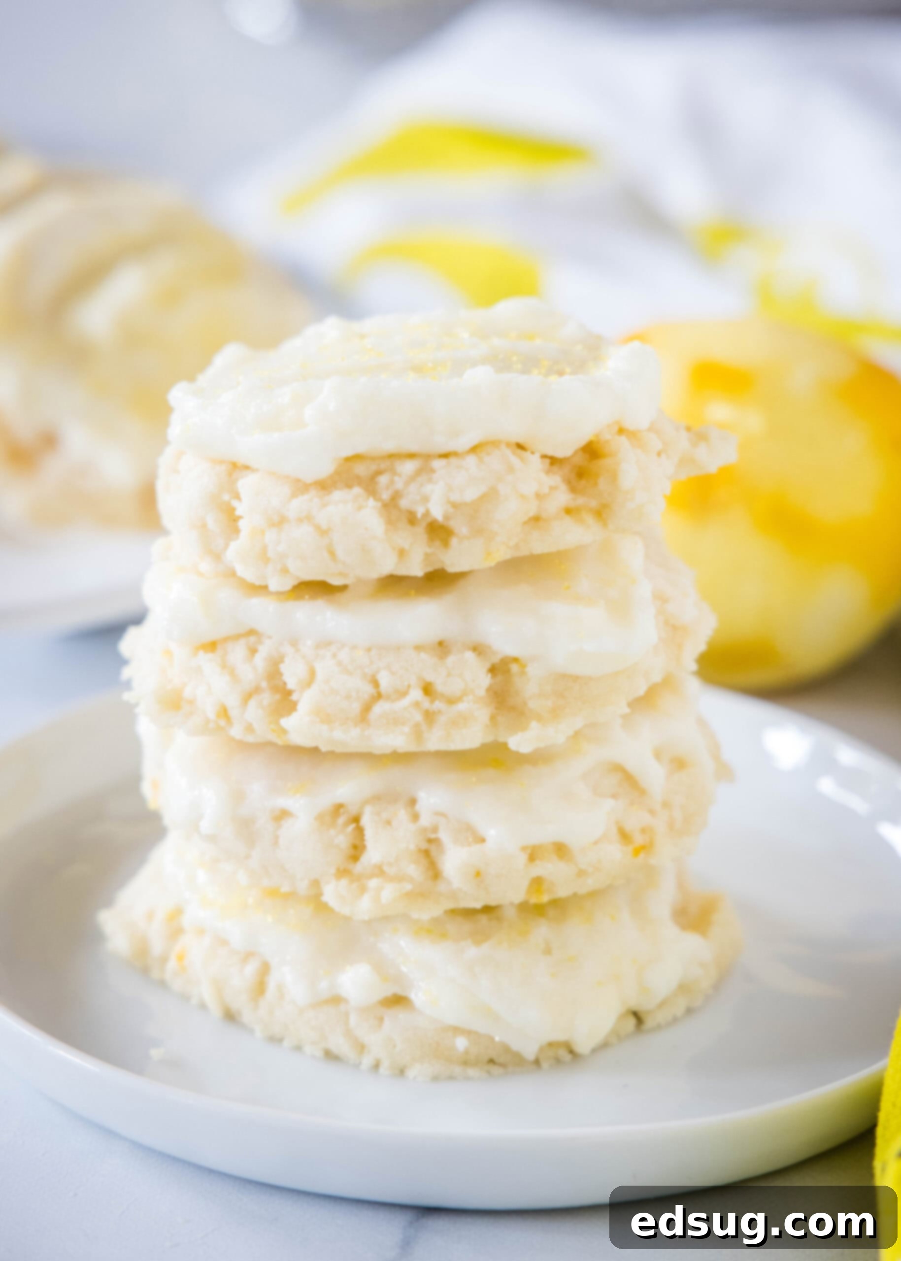 Soft lemon sugar cookies put a bright, zesty spin on a classic recipe! Made from real lemon juice and zest, topped with creamy lemon frosting.  Four beautifully frosted lemon sugar cookies stacked on a white plate with a zested lemon in the background, highlighting their soft texture and bright yellow frosting.