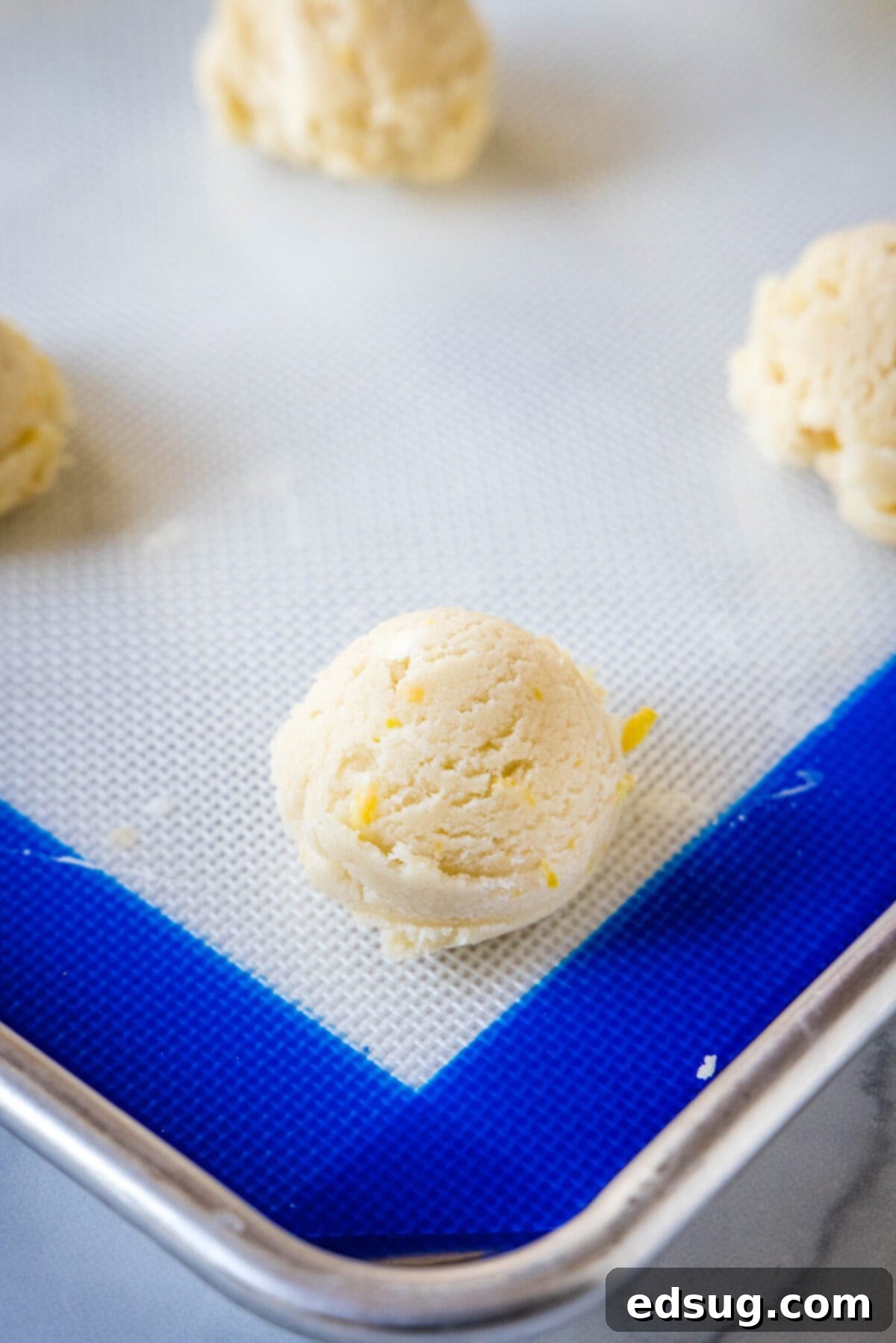 Soft lemon sugar cookies put a bright, zesty spin on a classic recipe! Made from real lemon juice and zest, topped with creamy lemon frosting.  Evenly portioned lemon sugar cookie dough balls, perfectly shaped and ready for baking, arranged on a silpat-lined baking sheet.