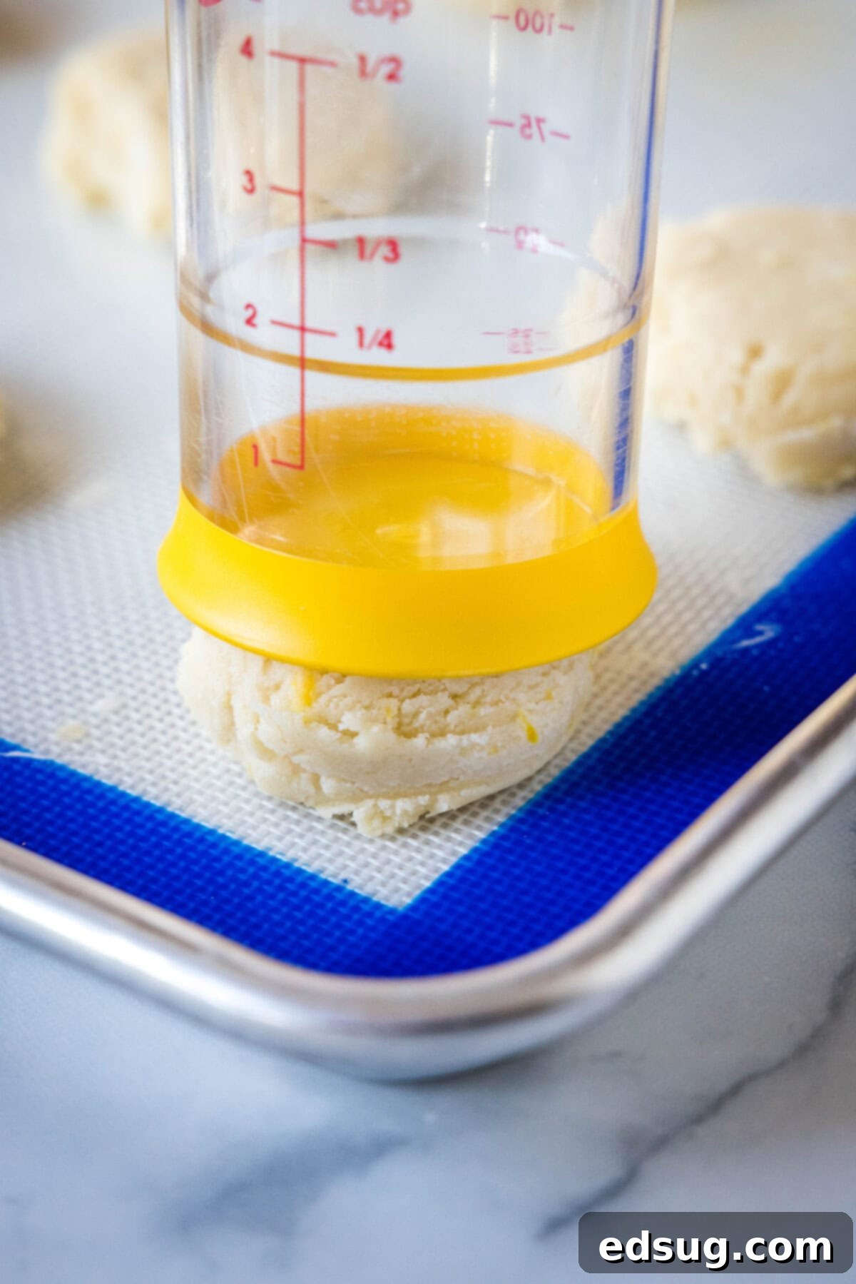Soft lemon sugar cookies put a bright, zesty spin on a classic recipe! Made from real lemon juice and zest, topped with creamy lemon frosting.  A measuring cup gently pressing down on the soft lemon cookie dough balls on a baking sheet, preparing them for baking to ensure an even shape.