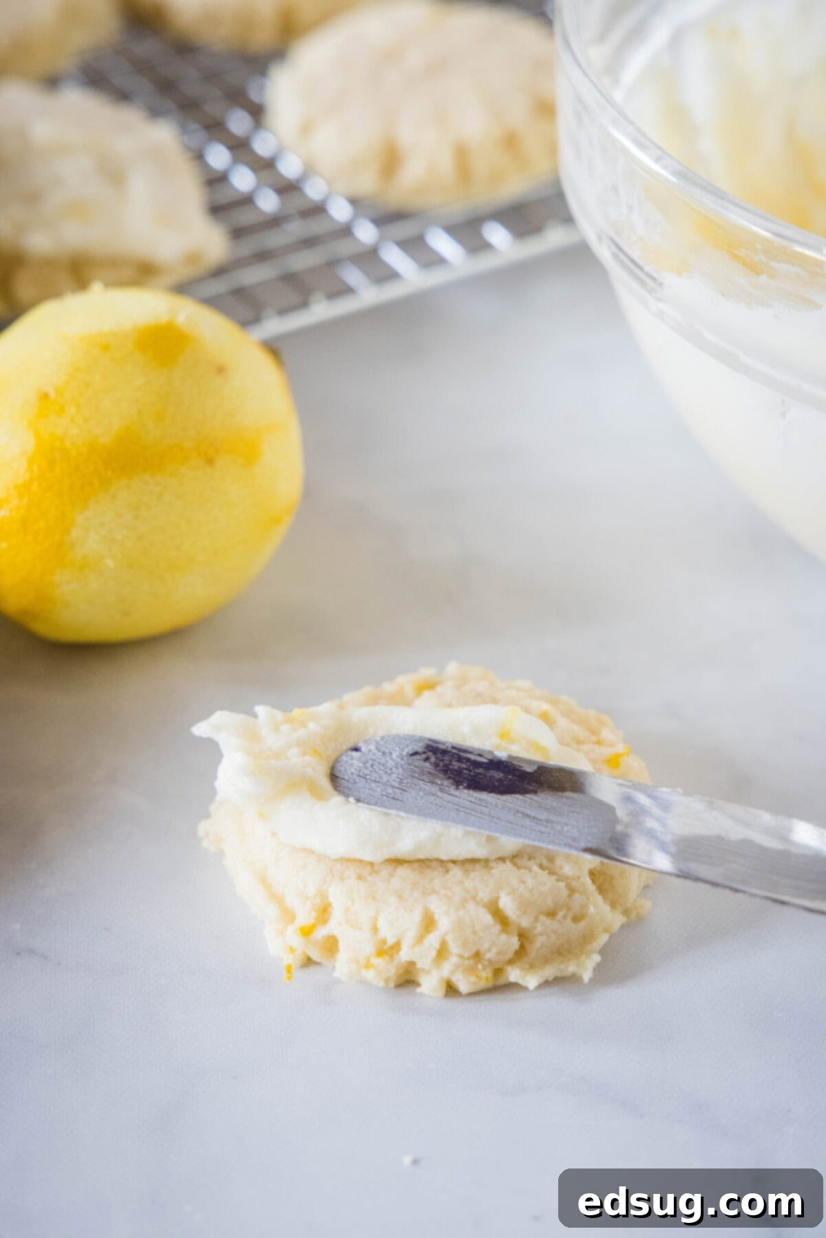 Soft lemon sugar cookies put a bright, zesty spin on a classic recipe! Made from real lemon juice and zest, topped with creamy lemon frosting.  A small spatula gracefully spreading creamy lemon frosting over the top of a freshly baked lemon sugar cookie, with more cookies cooling on a wire rack in the background.