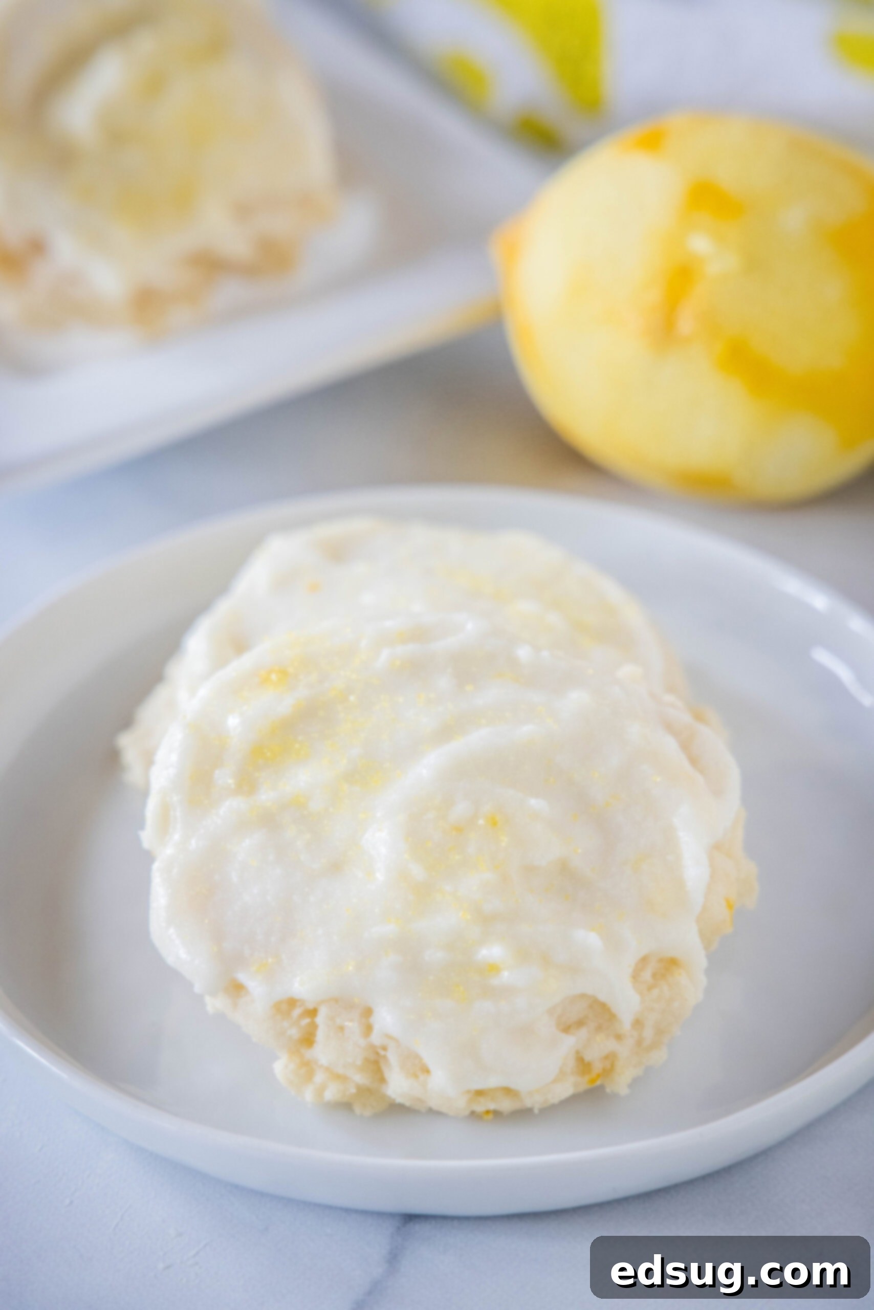 Soft lemon sugar cookies put a bright, zesty spin on a classic recipe! Made from real lemon juice and zest, topped with creamy lemon frosting.  Two perfectly frosted lemon sugar cookies, adorned with yellow sprinkles, resting on a pristine white plate with a zested lemon in the background, ready to be enjoyed.