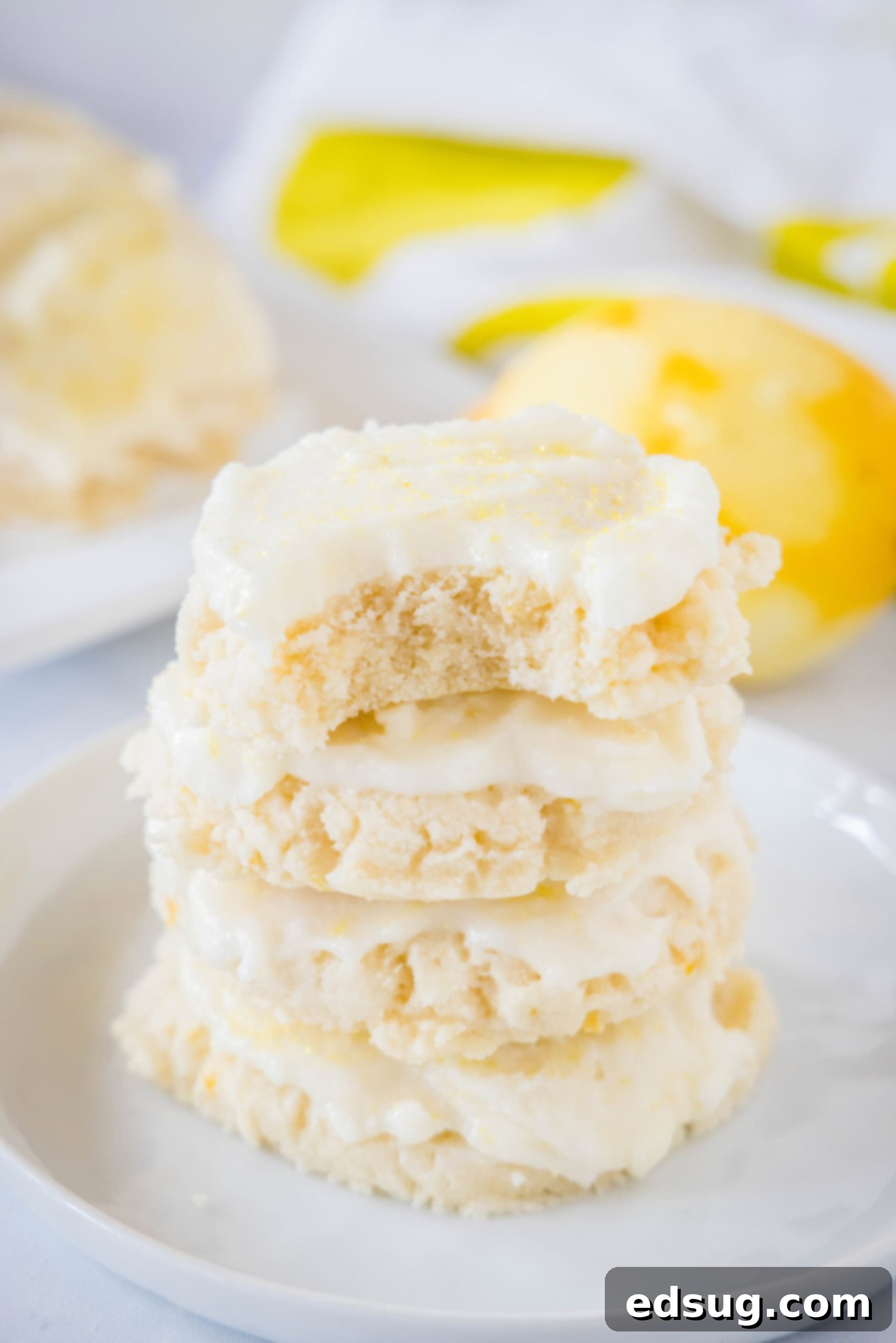Soft lemon sugar cookies put a bright, zesty spin on a classic recipe! Made from real lemon juice and zest, topped with creamy lemon frosting.  A stack of four frosted lemon sugar cookies on a white plate, with a bite taken out of the top cookie, revealing its soft, chewy interior and bright lemon flavor.