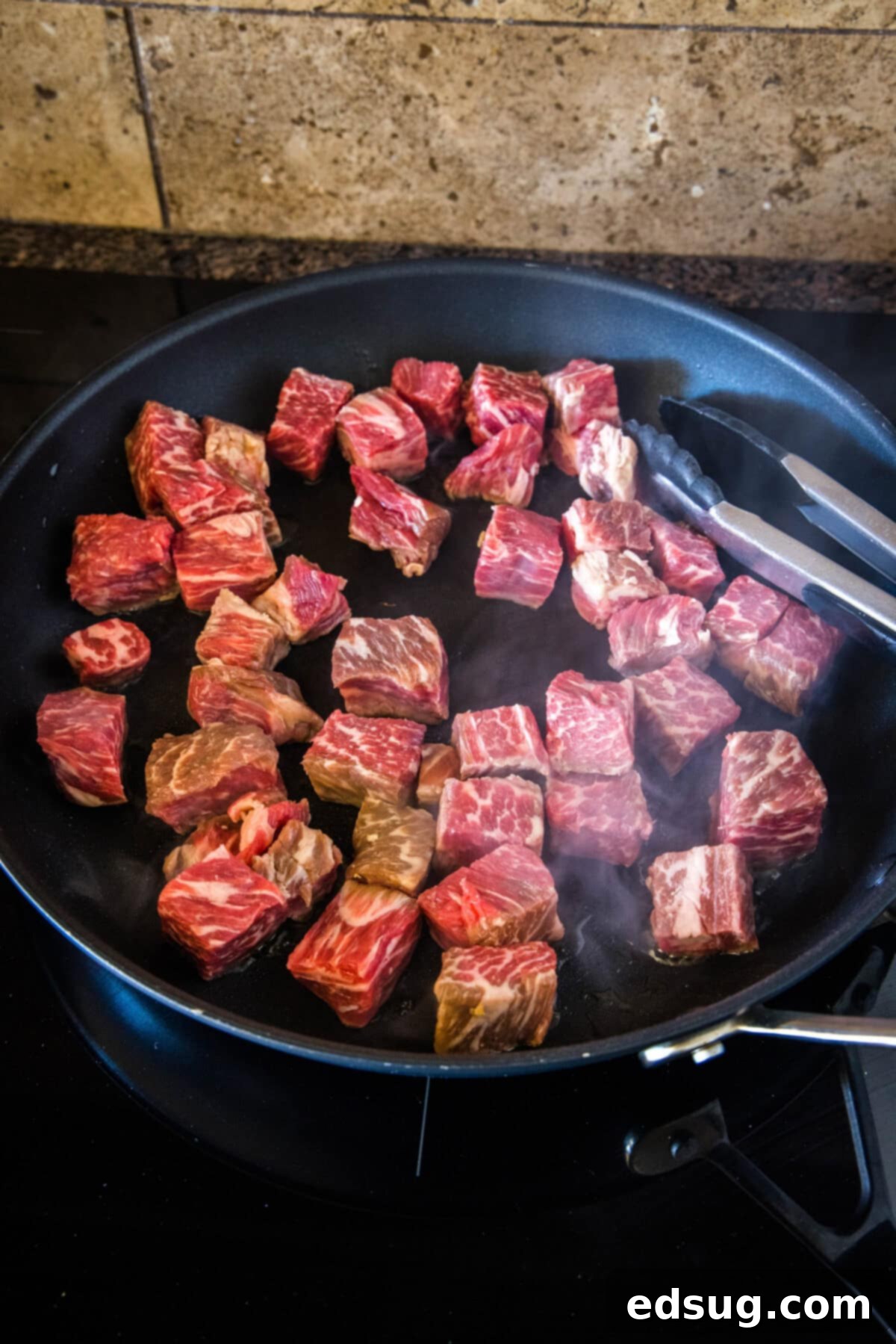 Steak bites searing in a skillet with a set of tongs.