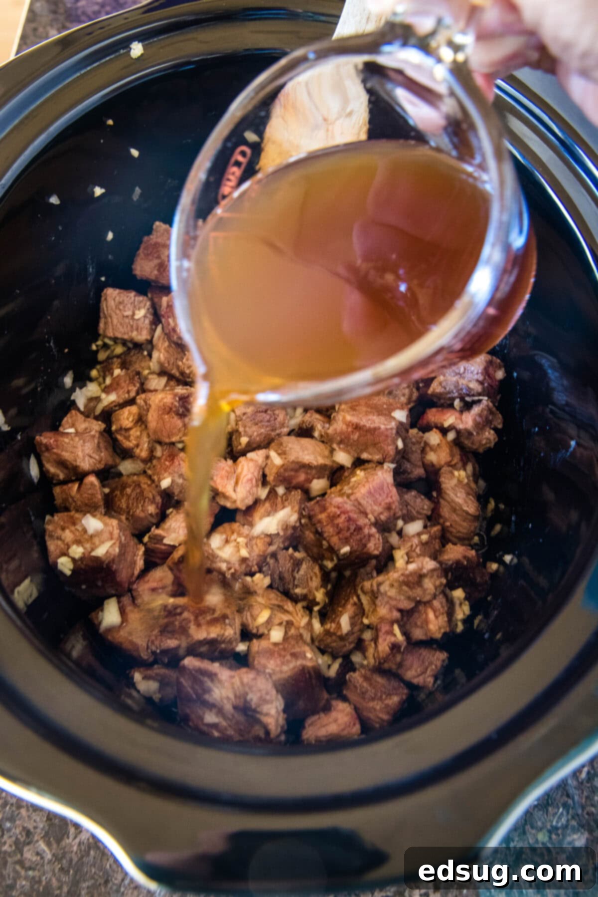 Beef broth being poured over steak bites ingredients in the crockpot.