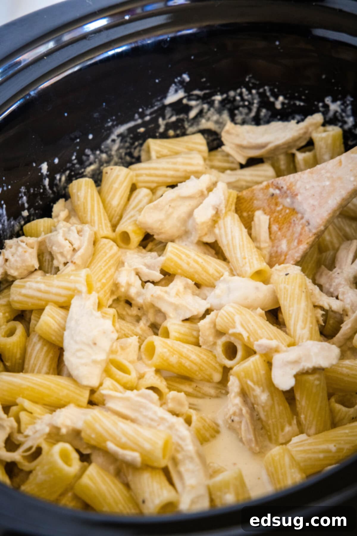 Shredded chicken and cooked pasta being stirred into alfredo sauce inside the slow cooker.