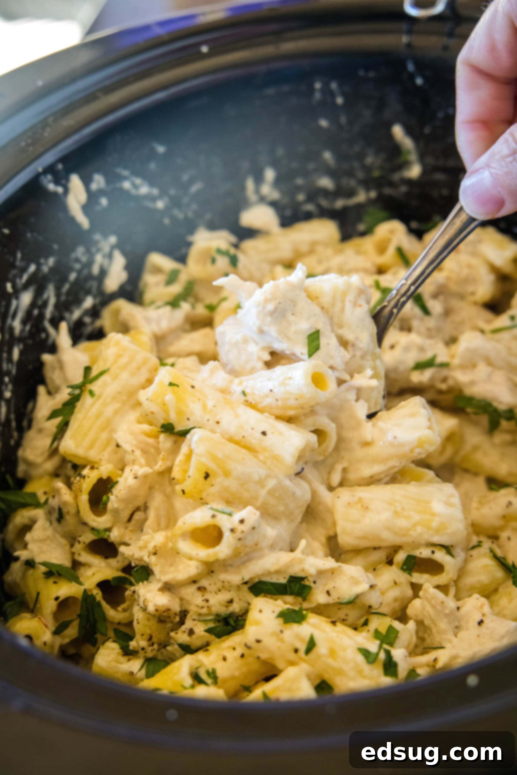 A hand using a spoon to stir chicken alfredo inside the crock pot.