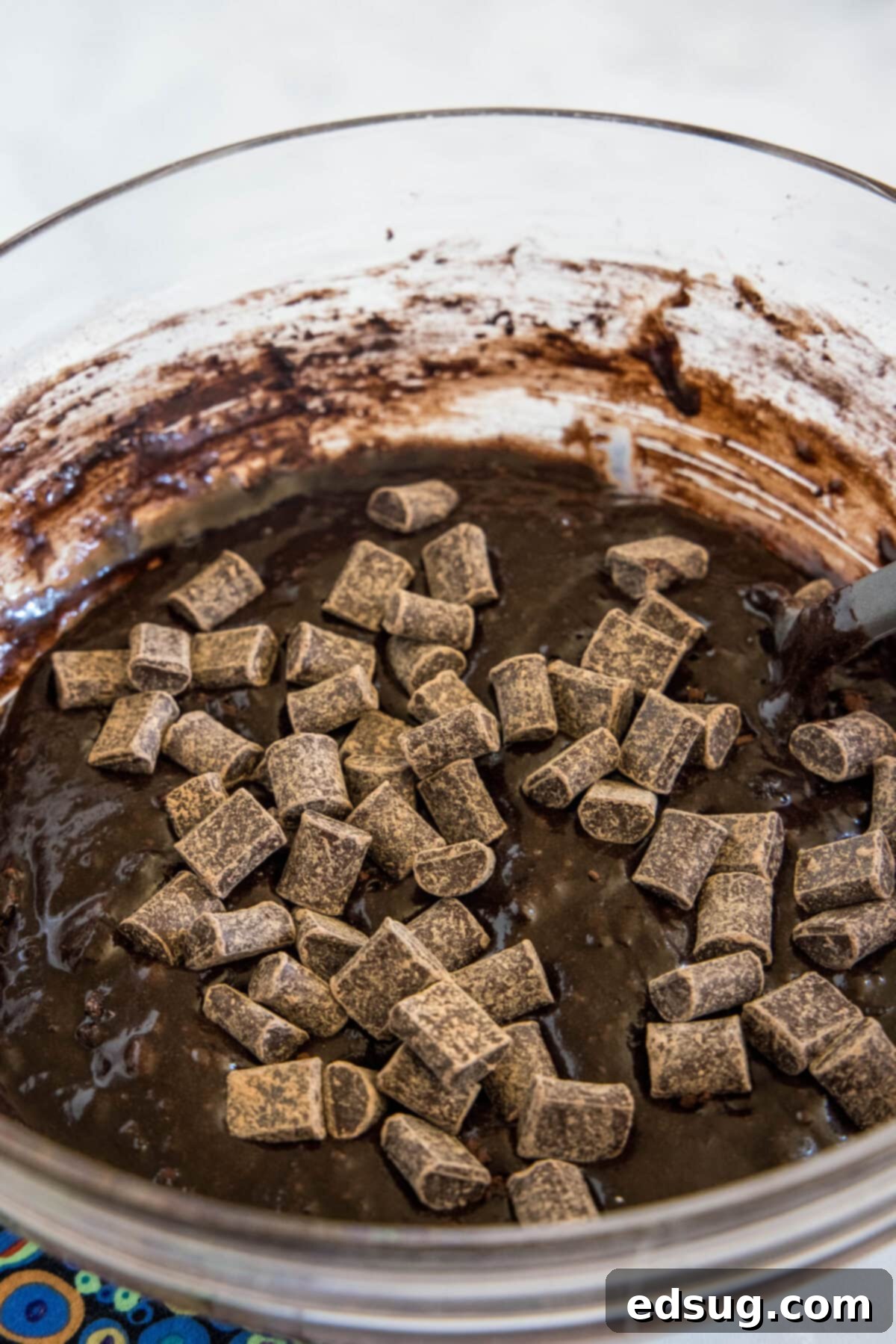 Make thick and chewy, fudgy double chocolate brownies with cocoa powder and chocolate chunks, for loads of melty chocolate in every bite! Generous chocolate chunks are folded into a rich, dark brownie batter within a glass mixing bowl, ensuring even distribution of melty chocolate throughout the fudgy mixture. The batter is thick and luscious.