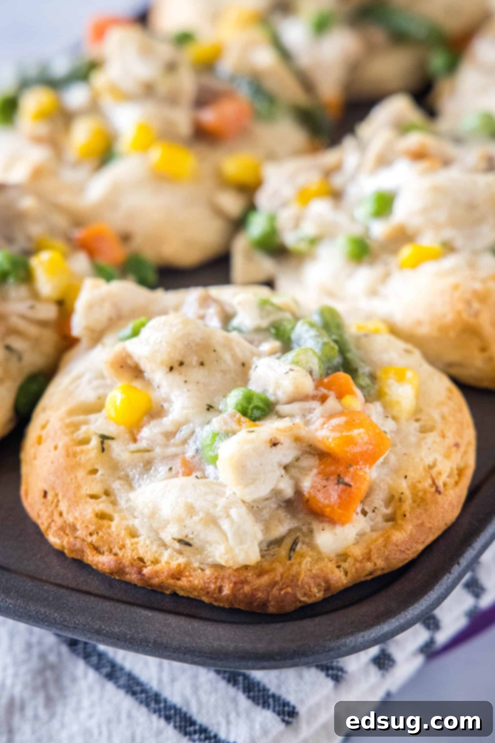 These mini chicken pot pies have creamy pot pie filling baked in a flaky biscuit crust. All the flavors of the original in bite-sized form! Close up of a mini chicken pot pie in a muffin tin with more mini pies in the background, showing golden crust and creamy filling.