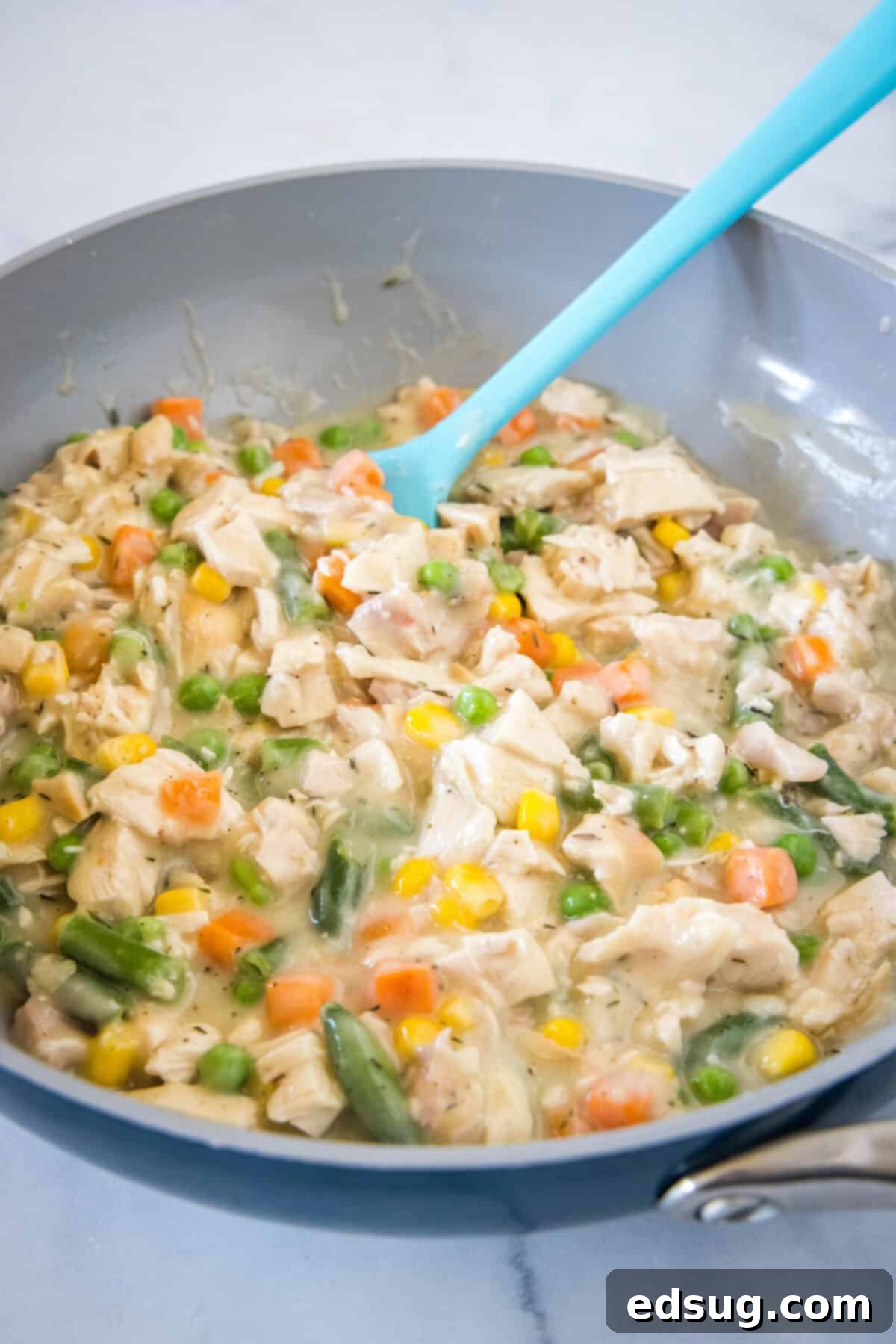 These mini chicken pot pies have creamy pot pie filling baked in a flaky biscuit crust. All the flavors of the original in bite-sized form! Chicken pot pie filling with cooked chicken and mixed vegetables being stirred in a skillet with a blue rubber spatula.