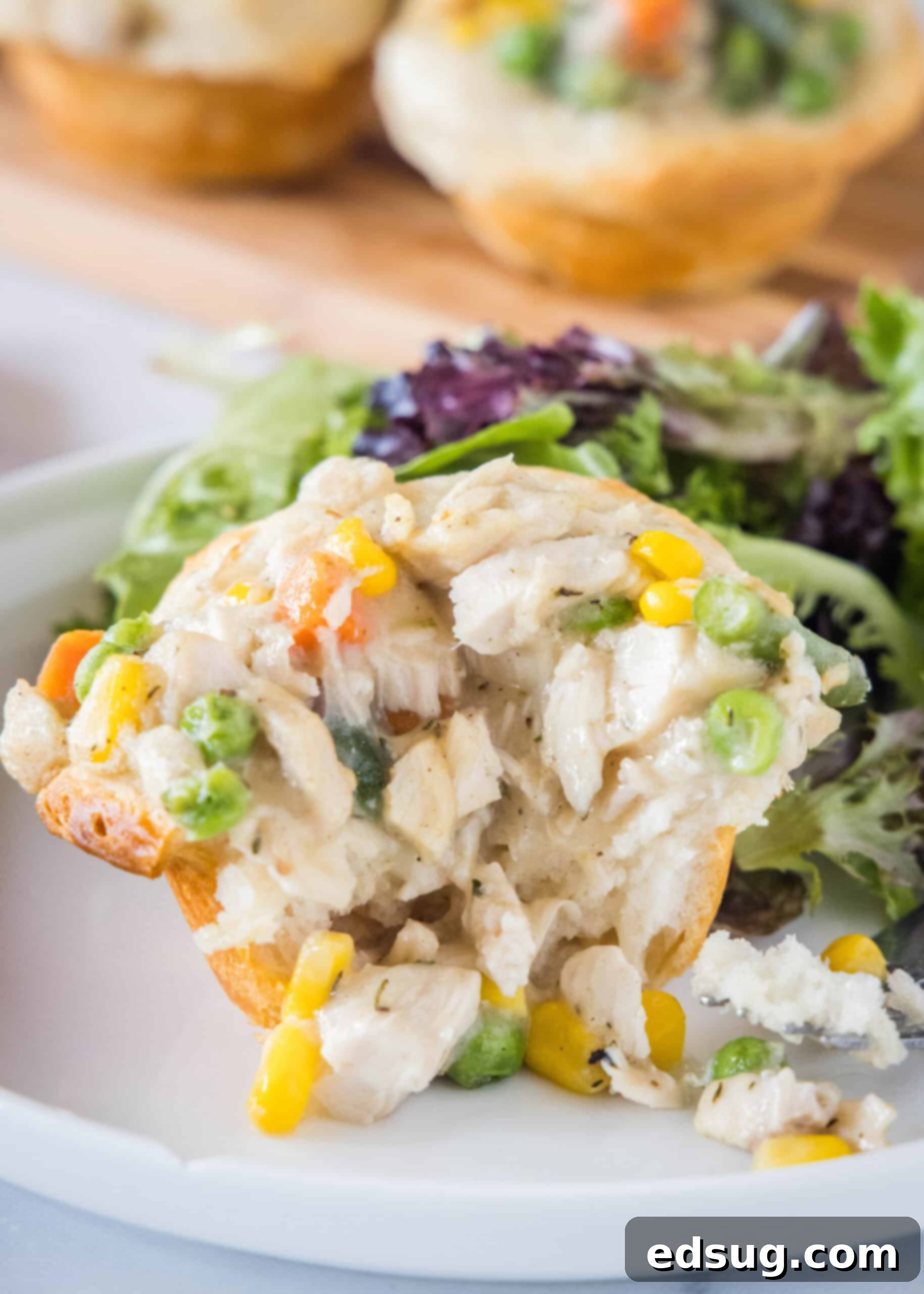 These mini chicken pot pies have creamy pot pie filling baked in a flaky biscuit crust. All the flavors of the original in bite-sized form! A mini chicken pot pie with a bite missing, showcasing the creamy filling and tender chicken, placed on a plate with a green salad in the background.