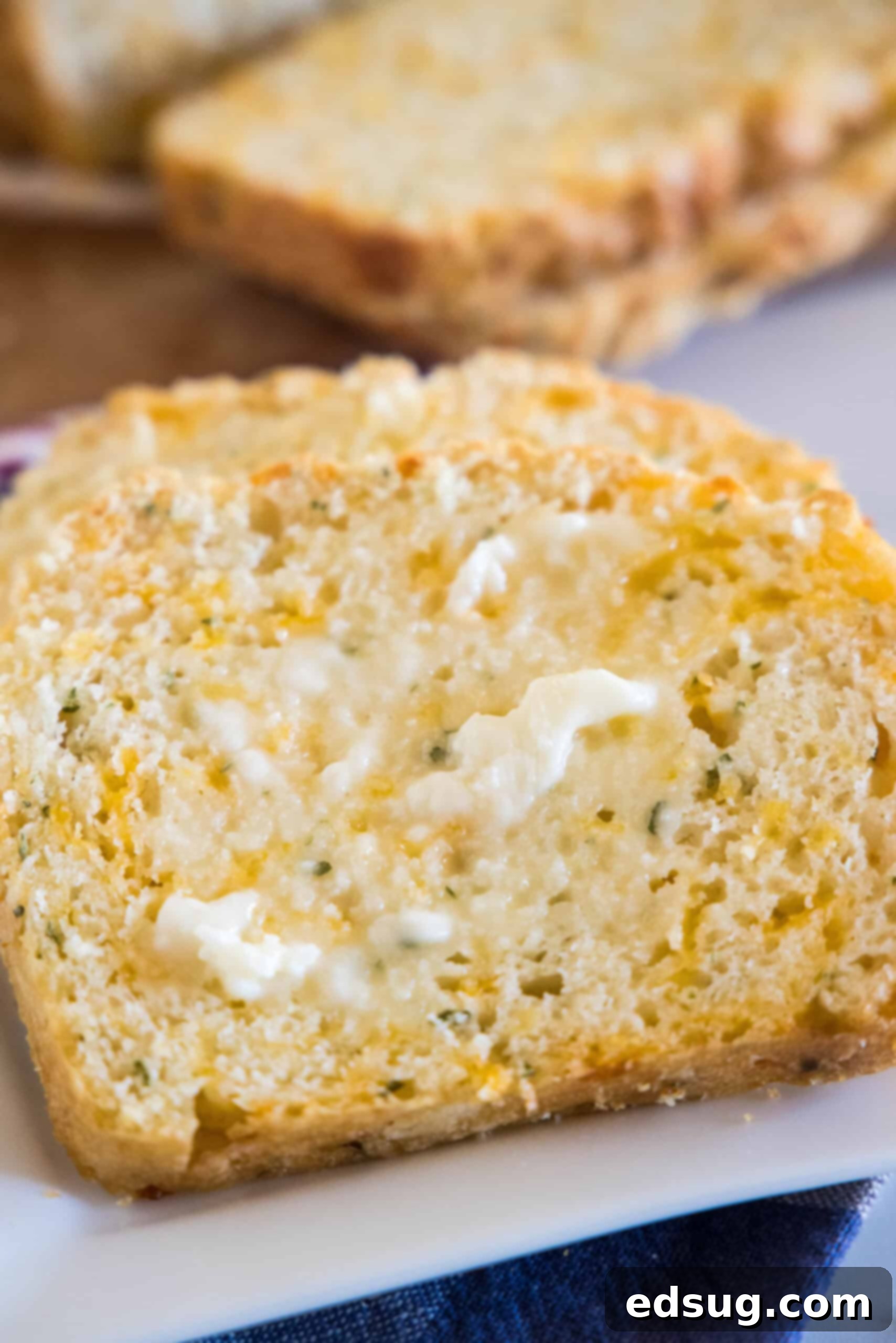 This cheesy quick bread recipe makes soft cheddar cheese bread filled with garlic and herbs. With no yeast, it's perfect for everyday baking! Close up of a thick slice of golden-brown cheesy quick bread, generously spread with butter, with more slices neatly stacked in the blurred background. The soft, airy crumb and visible specks of herbs are highlighted.