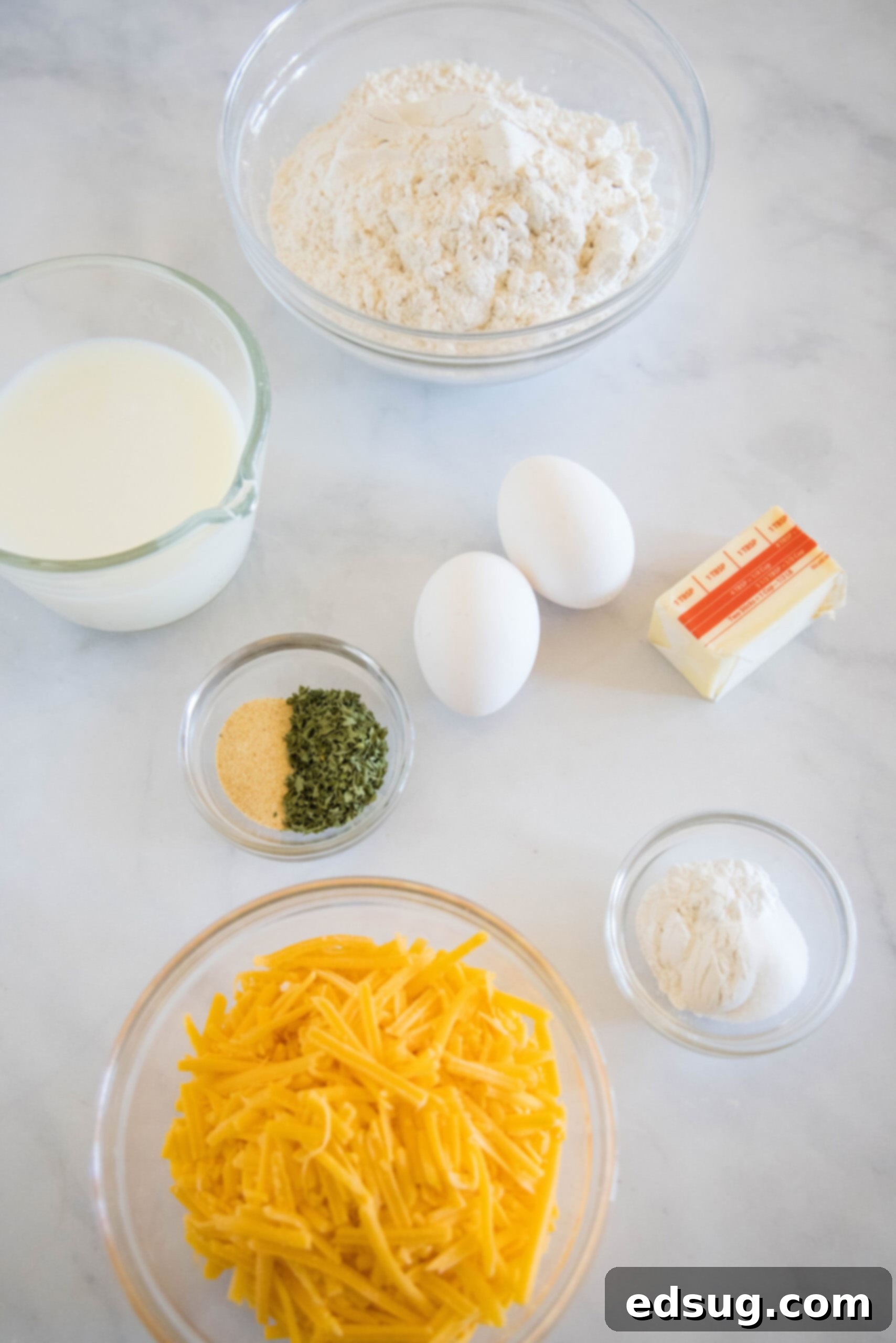 This cheesy quick bread recipe makes soft cheddar cheese bread filled with garlic and herbs. With no yeast, it's perfect for everyday baking! A collection of fresh ingredients for cheesy quick bread, including a bowl of flour, cubed butter, shredded cheddar cheese, whole eggs, a glass of milk, and small bowls of garlic powder and dried parsley, arranged neatly on a wooden countertop.