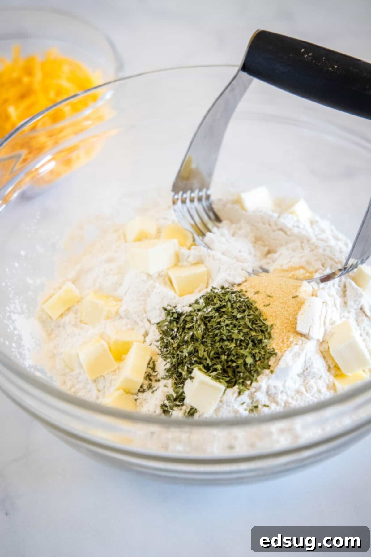 This cheesy quick bread recipe makes soft cheddar cheese bread filled with garlic and herbs. With no yeast, it's perfect for everyday baking! Cubed cold butter and dried herbs being added to a bowl of all-purpose flour, positioned next to a pastry cutter, illustrating the start of the 'cut in butter' step for the cheesy quick bread recipe.