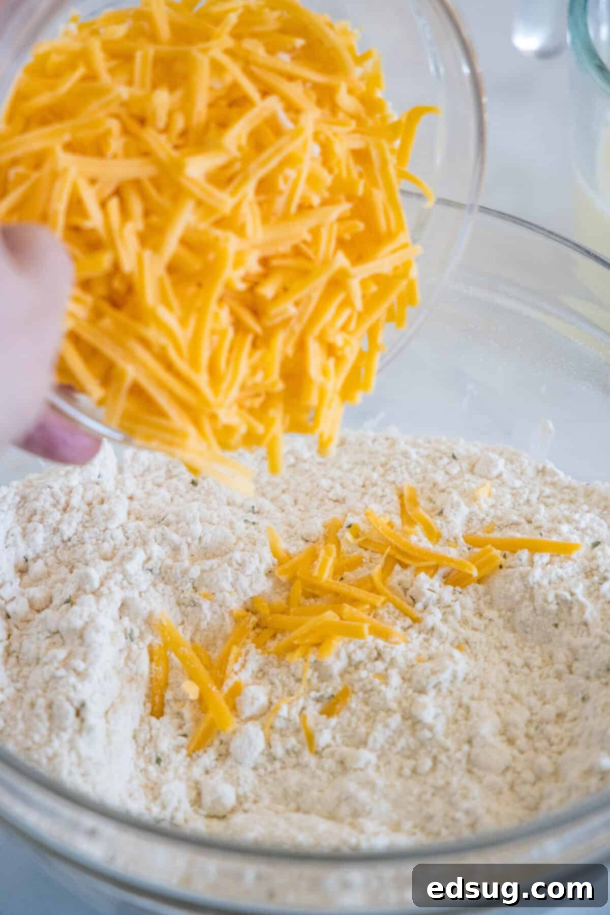 This cheesy quick bread recipe makes soft cheddar cheese bread filled with garlic and herbs. With no yeast, it's perfect for everyday baking! A generous amount of shredded cheddar cheese being added to a bowl containing a flour and butter crumble mixture, ready for mixing into the cheesy quick bread batter.