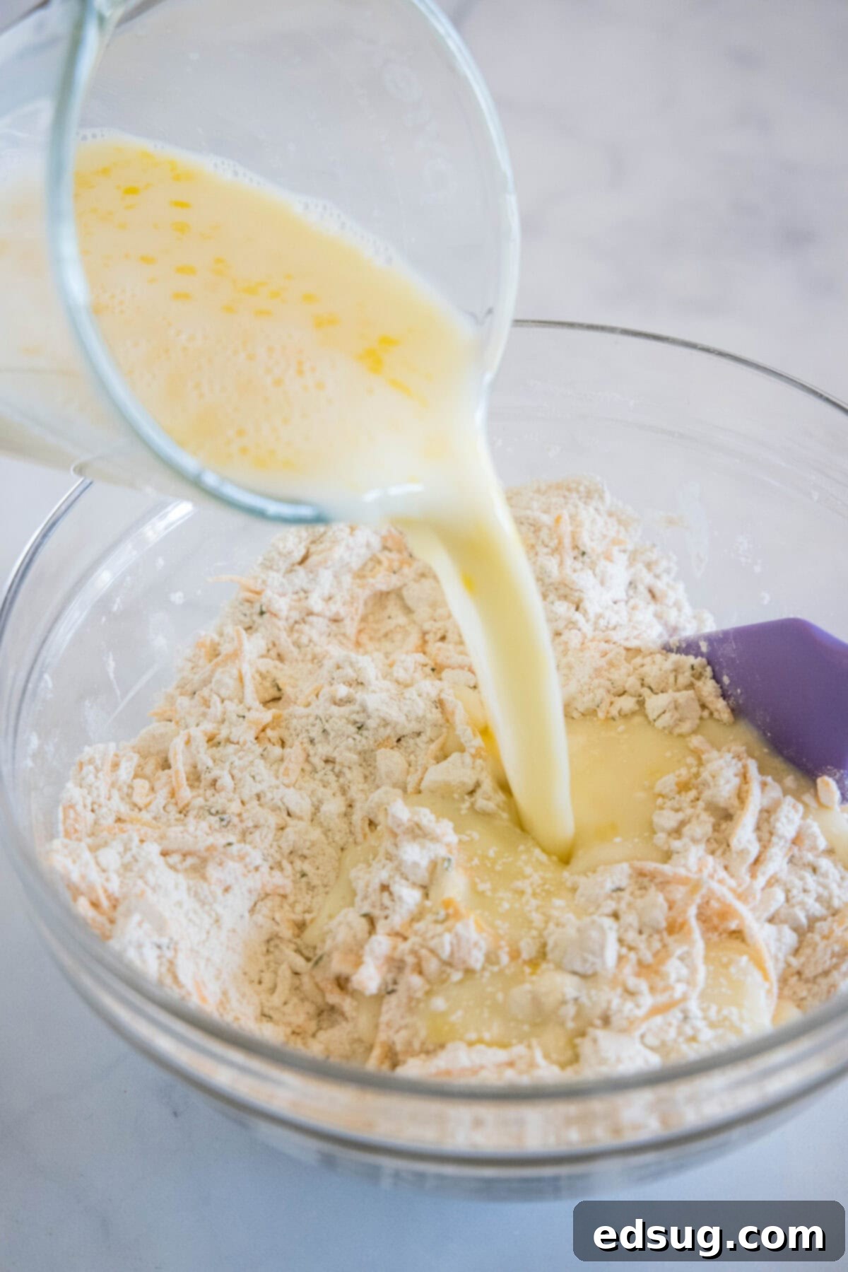 This cheesy quick bread recipe makes soft cheddar cheese bread filled with garlic and herbs. With no yeast, it's perfect for everyday baking! A stream of whisked wet ingredients (eggs and milk) being poured into a bowl containing the dry, cheesy bread ingredients, illustrating the process of combining for the cheesy quick bread recipe.