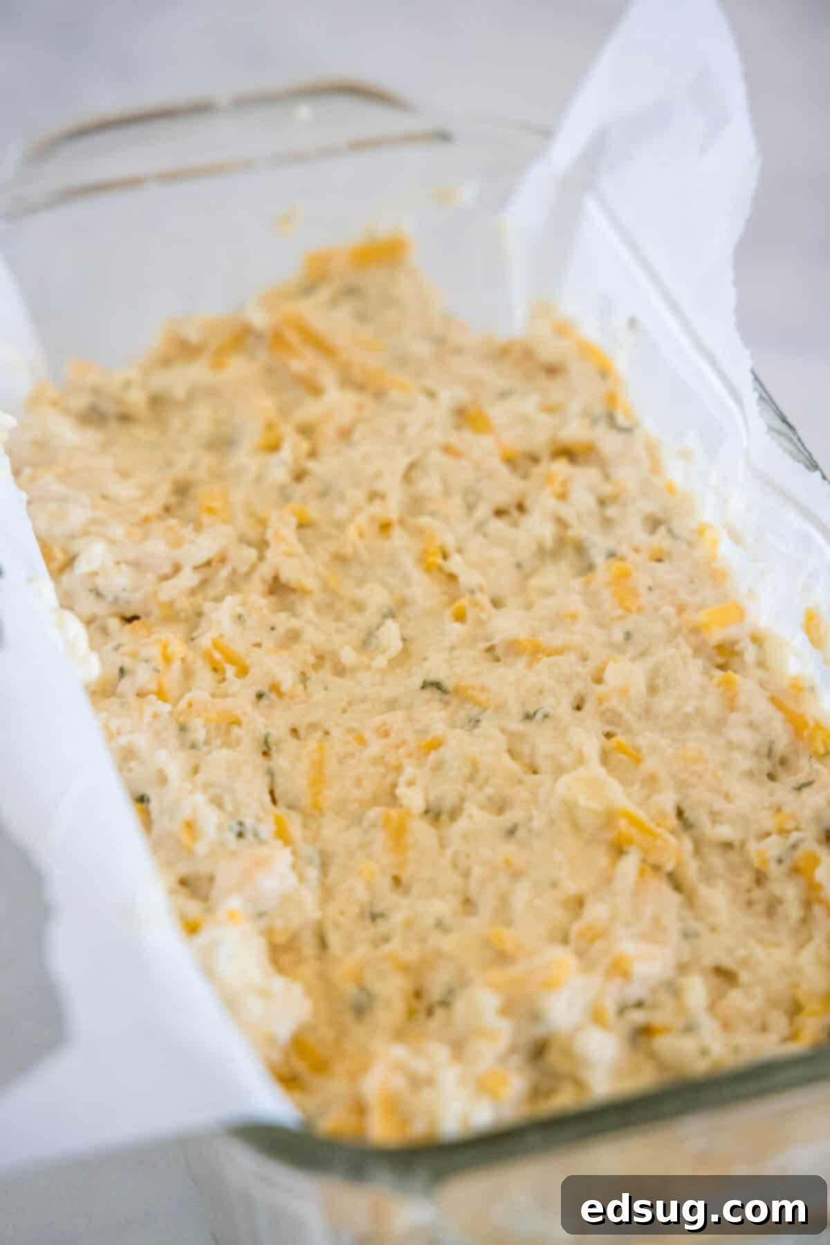 This cheesy quick bread recipe makes soft cheddar cheese bread filled with garlic and herbs. With no yeast, it's perfect for everyday baking! The finished cheesy bread batter being carefully added to a parchment-lined glass loaf pan, preparing it for baking to create a delicious, homemade loaf.