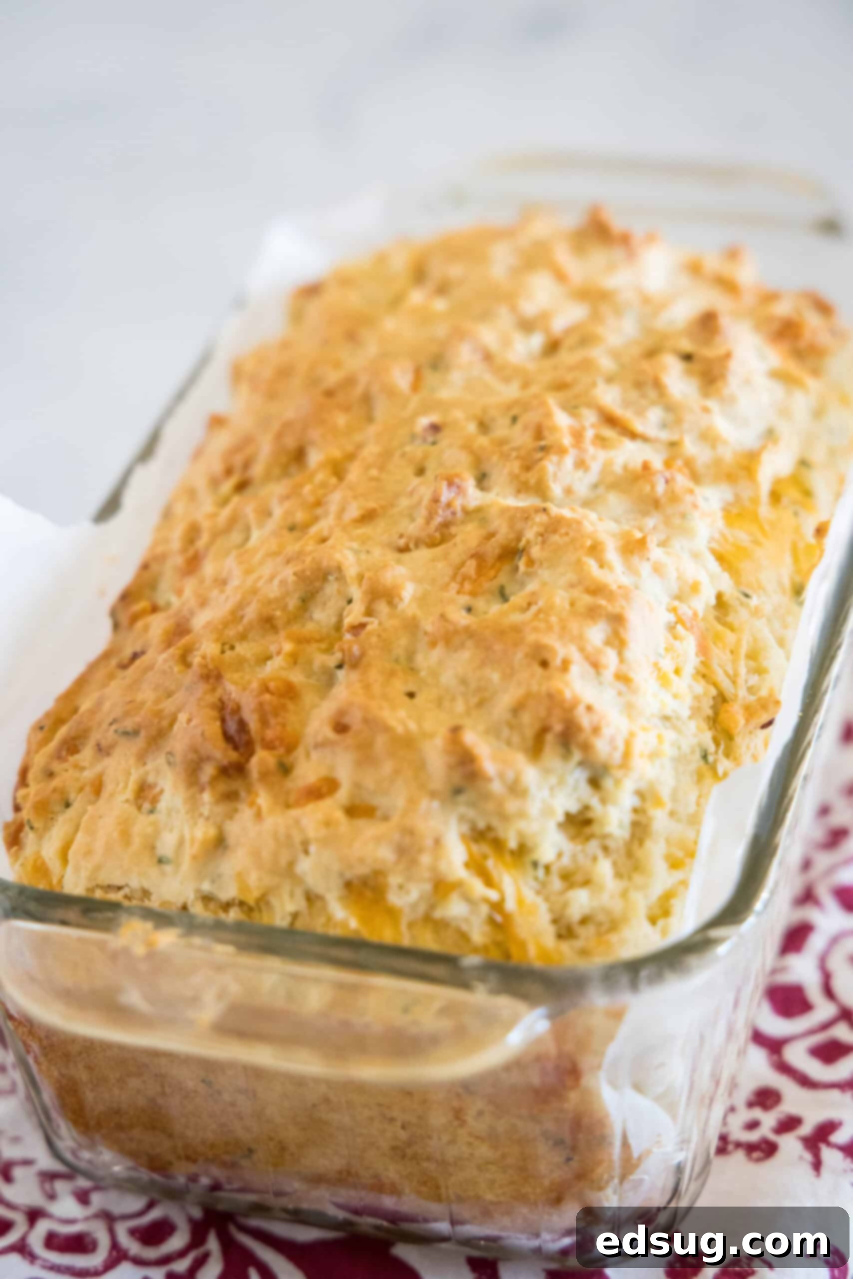 This cheesy quick bread recipe makes soft cheddar cheese bread filled with garlic and herbs. With no yeast, it's perfect for everyday baking! A perfectly baked, golden-brown loaf of cheesy quick bread, still warm and resting inside its parchment-lined glass loaf pan, just out of the oven.