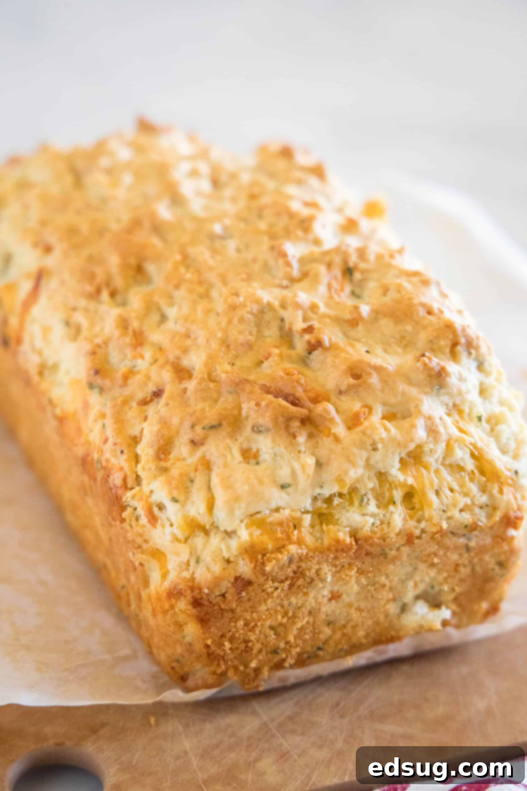 This cheesy quick bread recipe makes soft cheddar cheese bread filled with garlic and herbs. With no yeast, it's perfect for everyday baking! A freshly baked, golden-brown loaf of cheesy quick bread rests on a sheet of parchment paper on a rustic wooden cutting board, ready for slicing. The crust is perfectly browned, and the texture looks inviting.