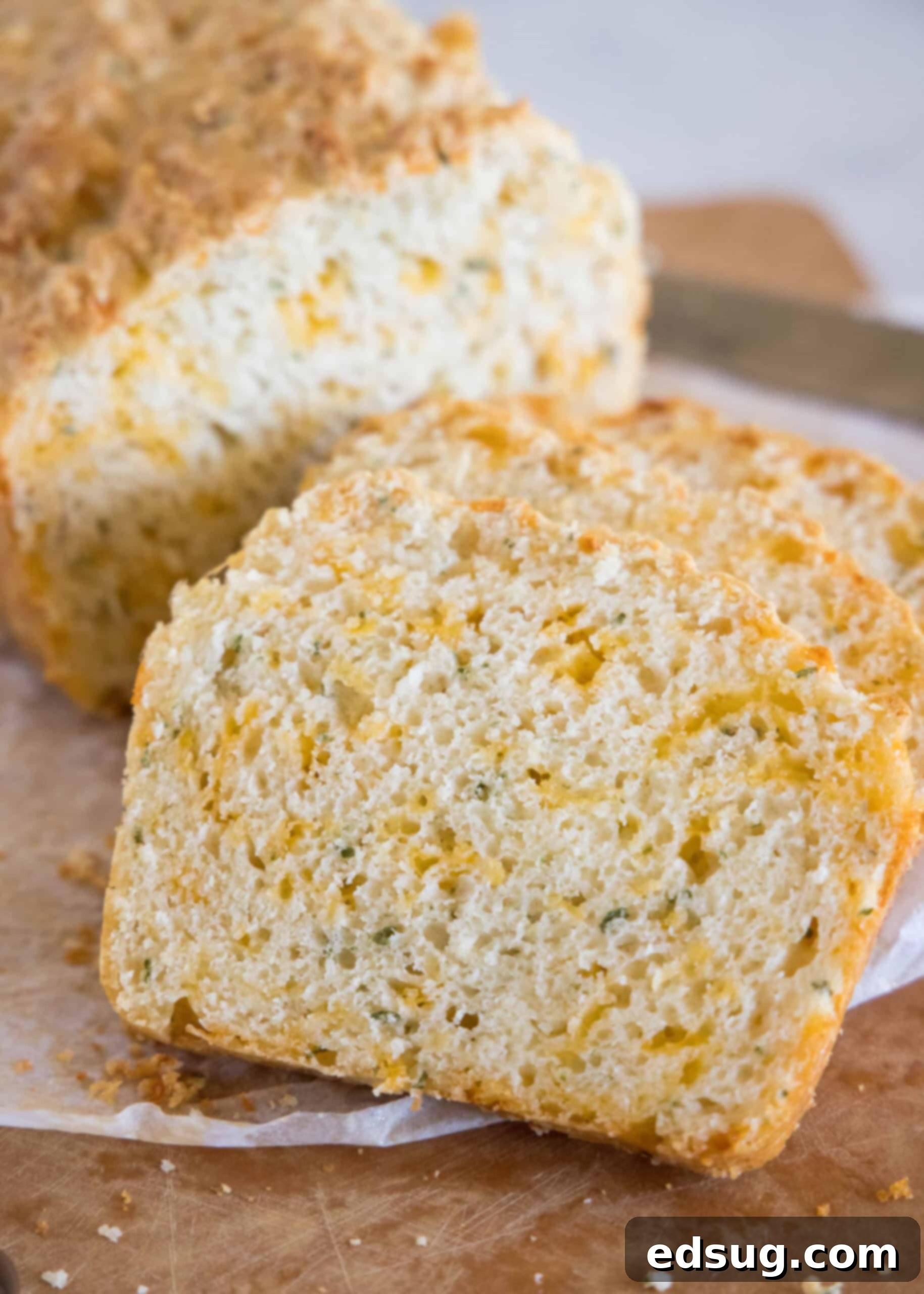 This cheesy quick bread recipe makes soft cheddar cheese bread filled with garlic and herbs. With no yeast, it's perfect for everyday baking! Several thick, golden-brown slices of cheddar quick bread, freshly cut from the end of a loaf, are artfully arranged on parchment paper atop a rustic wooden cutting board, ready for serving.
