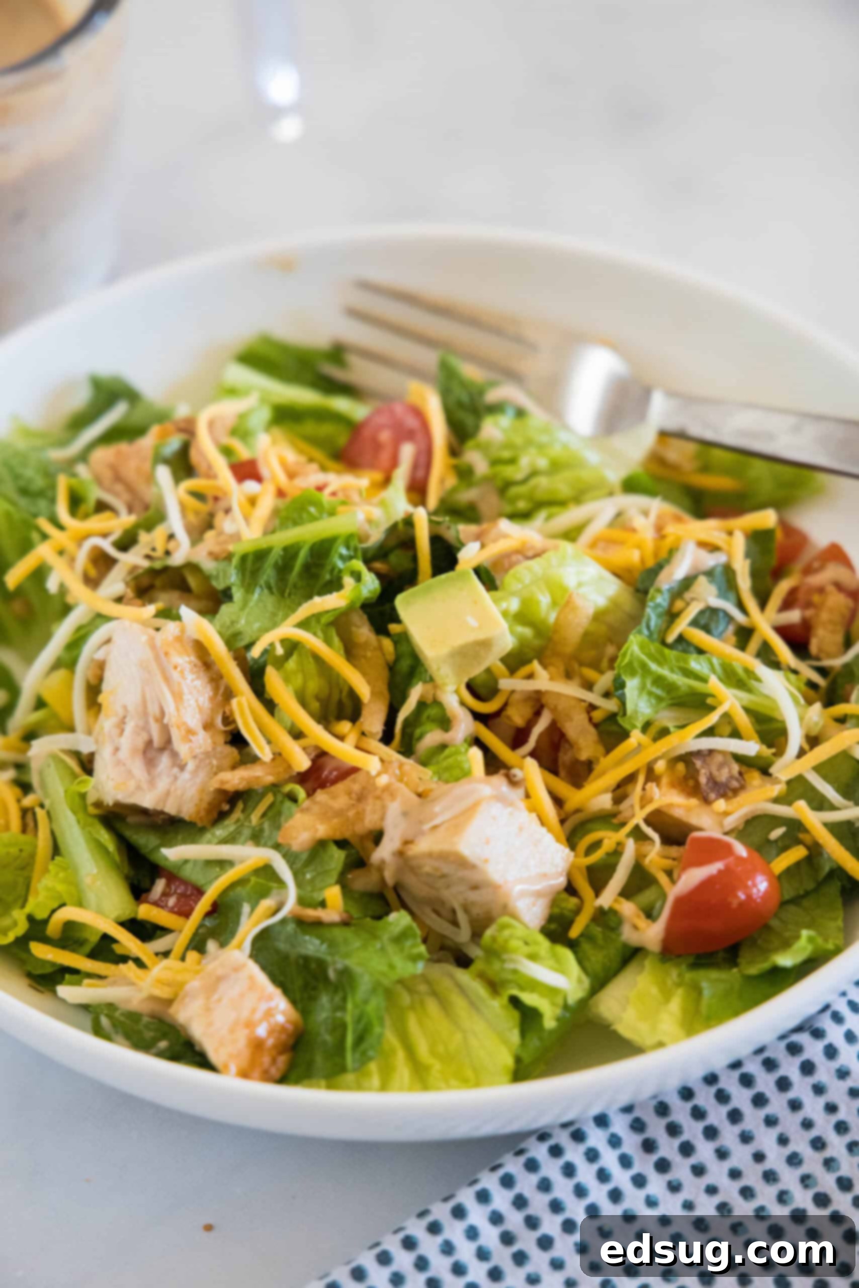 This BBQ chicken salad has juicy bites of sweet-smoky chicken tossed with tomatoes, avocado, and sweet corn in creamy barbecue ranch dressing. A large white bowl filled with a fresh and colorful BBQ chicken salad, featuring chopped chicken, lettuce, tomatoes, corn, avocado, and crispy fried onions, with a fork resting inside.