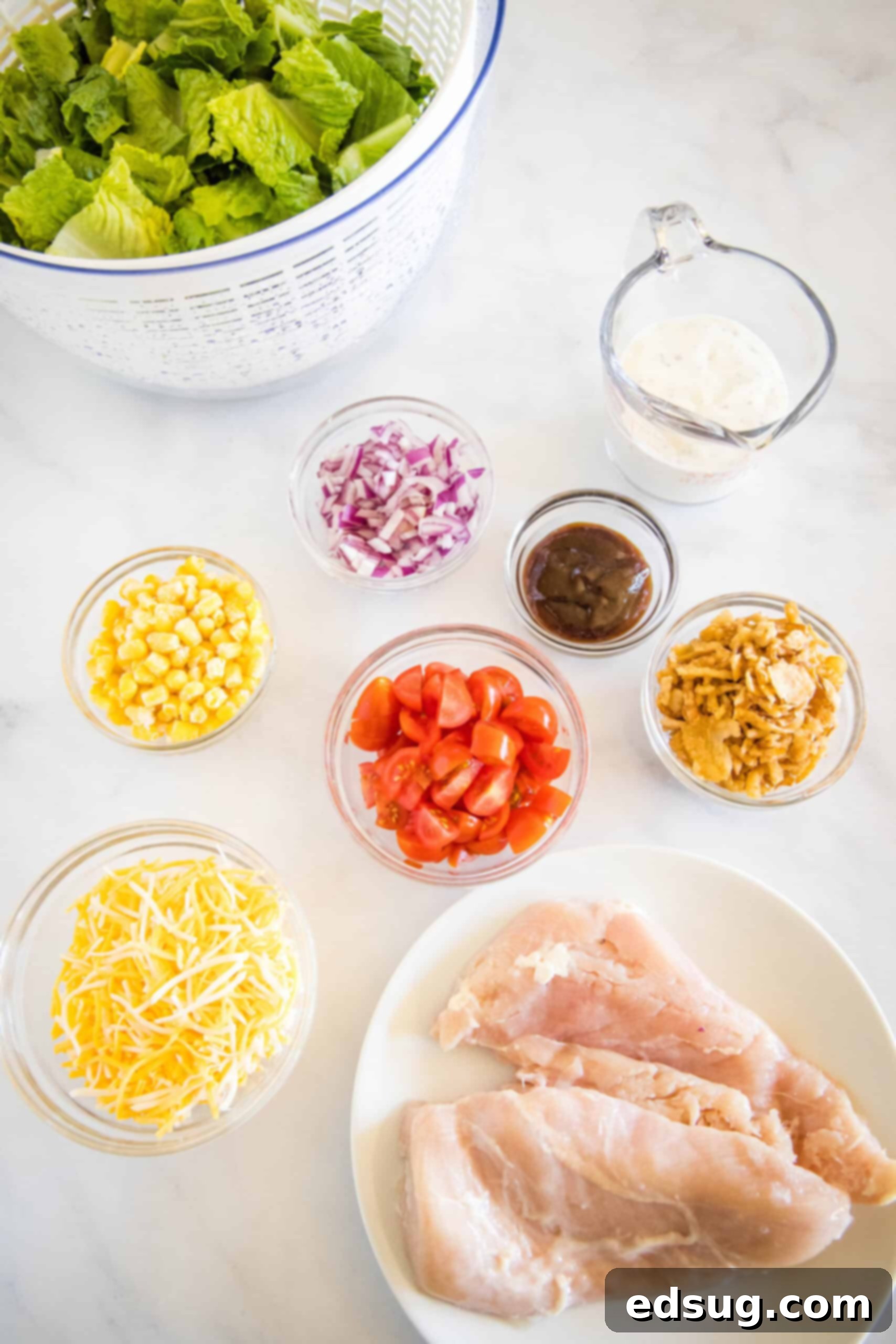 This BBQ chicken salad has juicy bites of sweet-smoky chicken tossed with tomatoes, avocado, and sweet corn in creamy barbecue ranch dressing. A flat lay image displaying all the fresh ingredients for a BBQ chicken salad, including chopped romaine, grape tomatoes, diced avocado, corn kernels, red onion, shredded Colby Jack cheese, cooked BBQ chicken, and a bowl of barbecue ranch dressing.